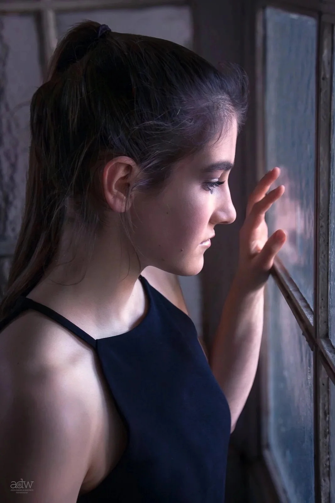 A young woman with dark hair in a ponytail looking out a window with her hand gently touching the glass.