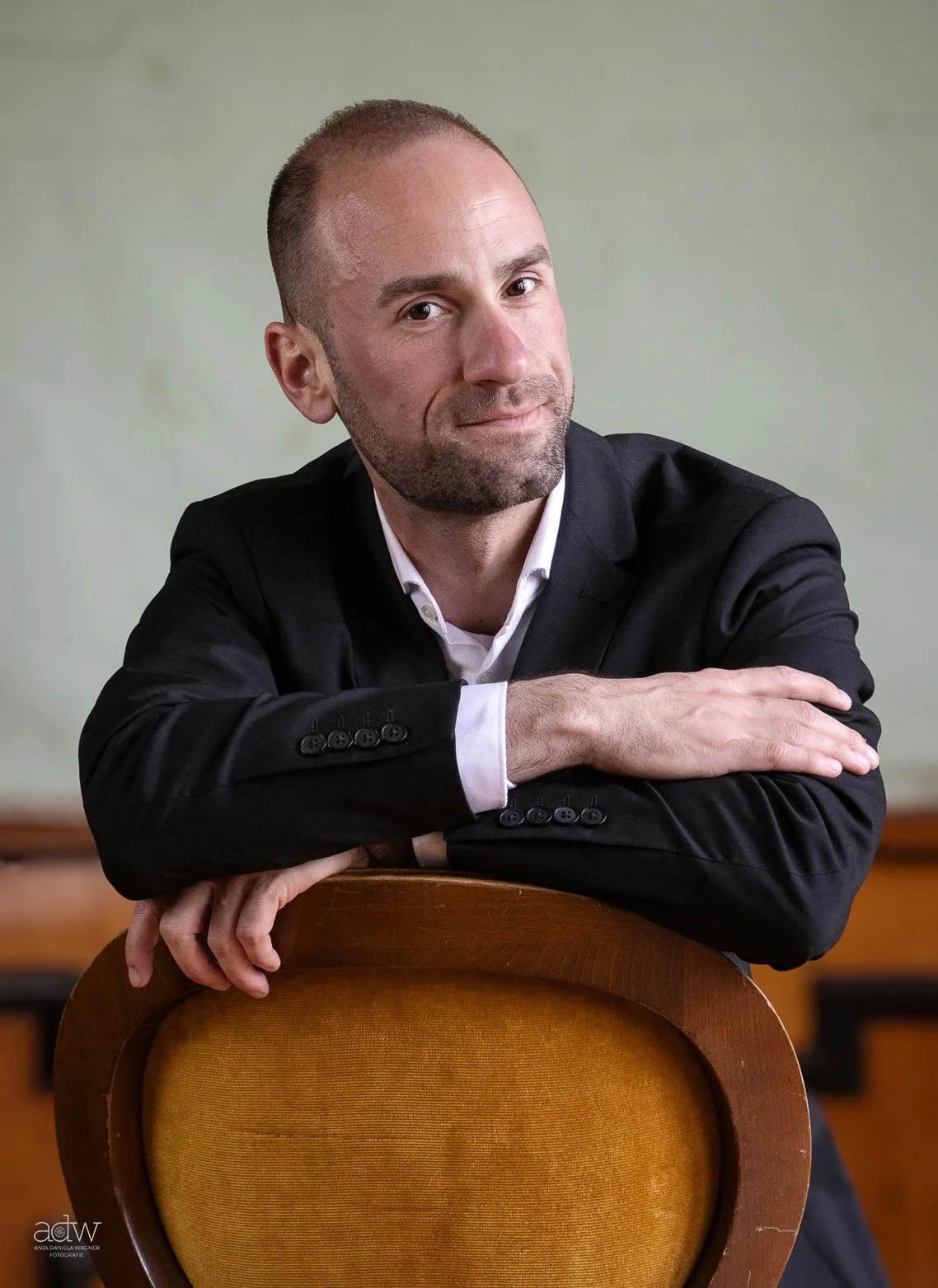 A man with short hair and a beard, dressed in a black suit and white shirt, leaning on an orange cushioned chair with a neutral background.