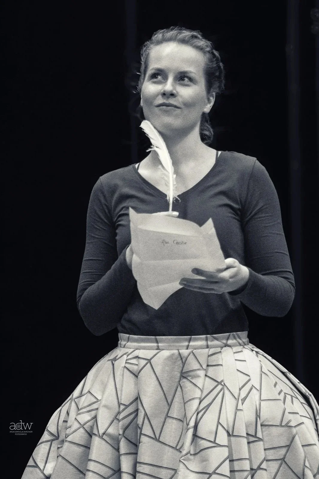 A woman with curly hair holding a white paper and a feather quill, wearing a dark long-sleeve top and a patterned skirt, standing against a dark background.