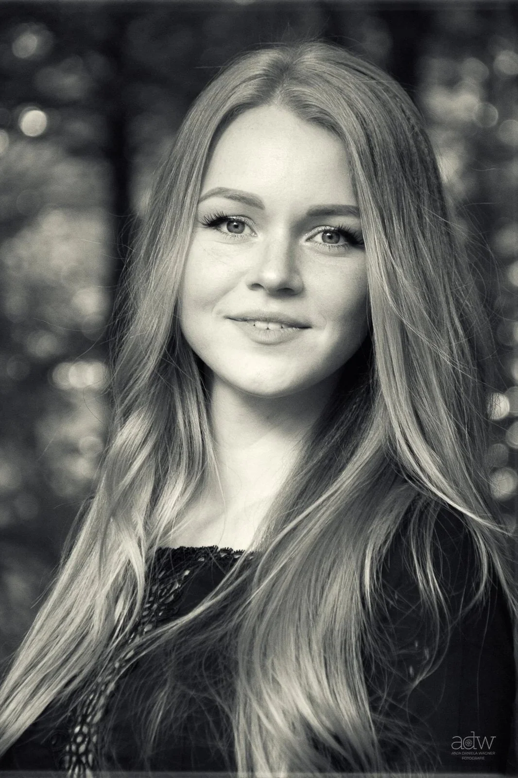 A black and white portrait of a young woman with long, wavy hair, smiling slightly, outdoors with blurred background.