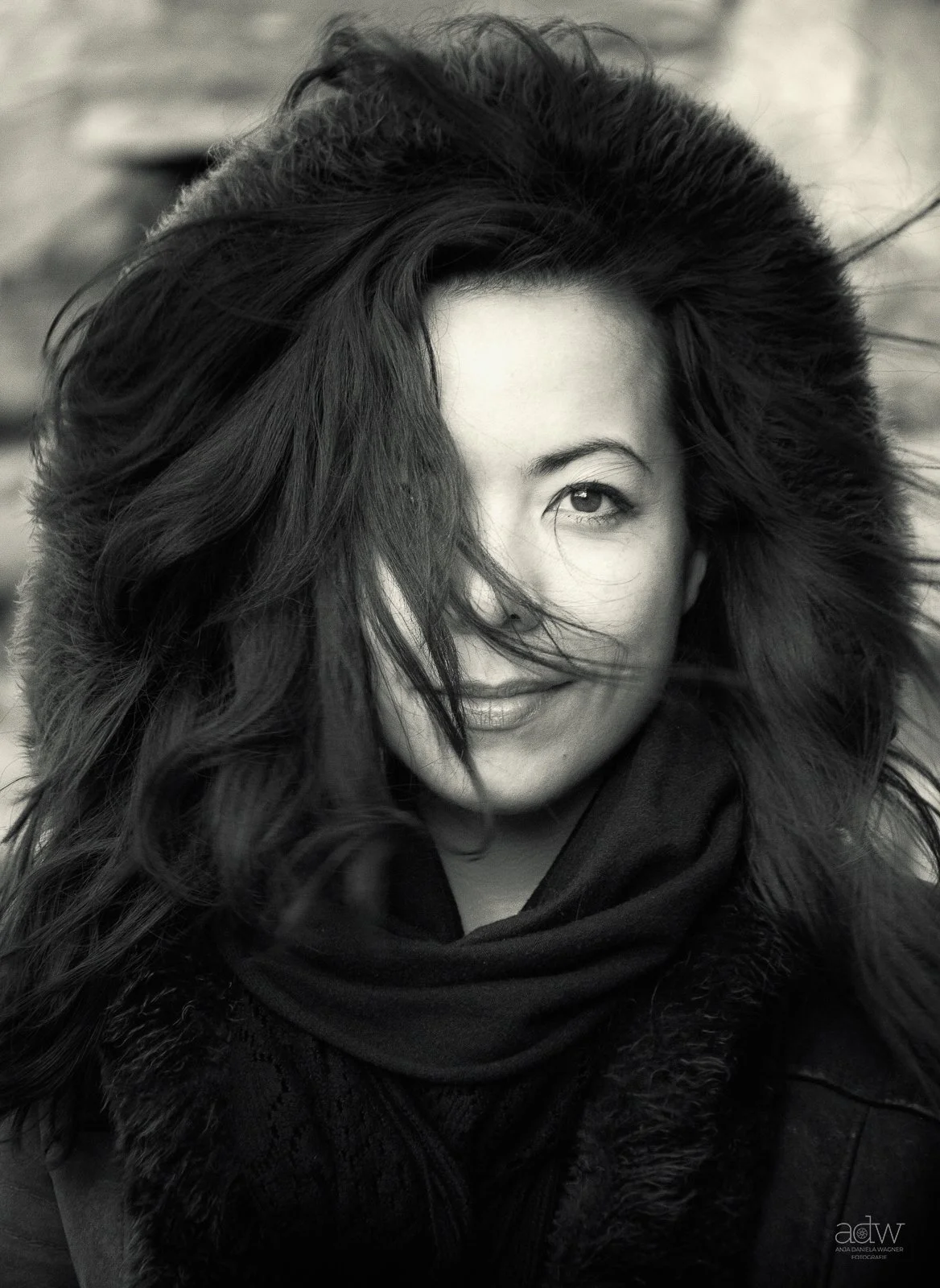 Black and white portrait of a woman smiling, with hair partially covering one eye, wearing a scarf and jacket.