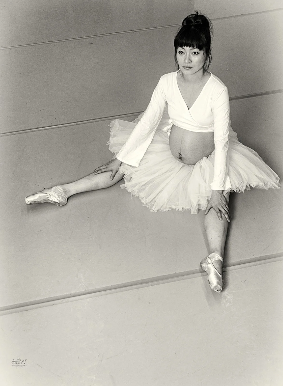 Black and white photo of a ballerina in a tutu practicing in front of a mirror, sitting on the floor with one leg extended and the other bent.