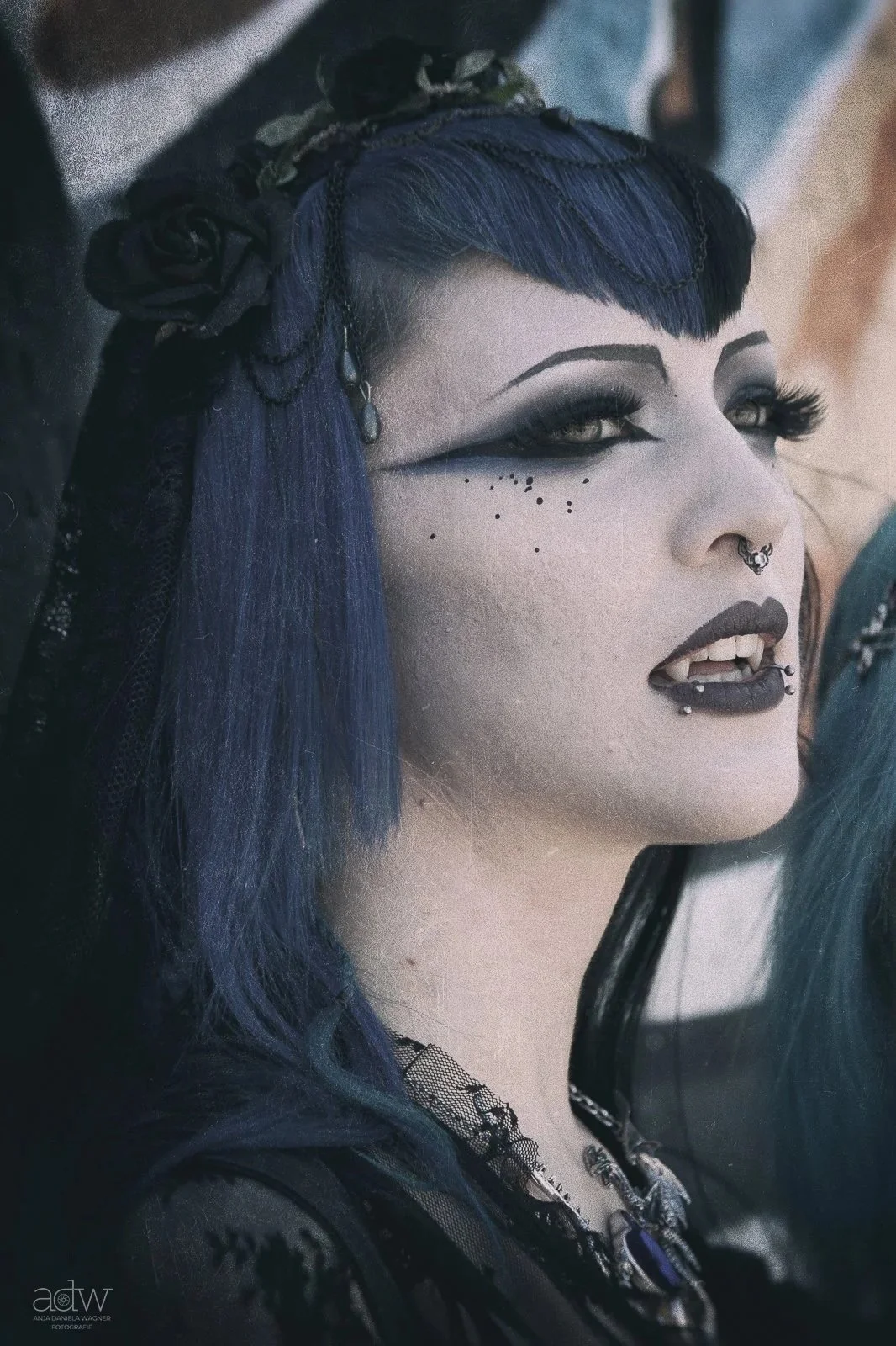 Close-up of a woman with pale skin, dark makeup, septum and lip piercings, black lipstick, and blue hair styled with black flowers and lace. She has dramatic eye makeup and freckles.