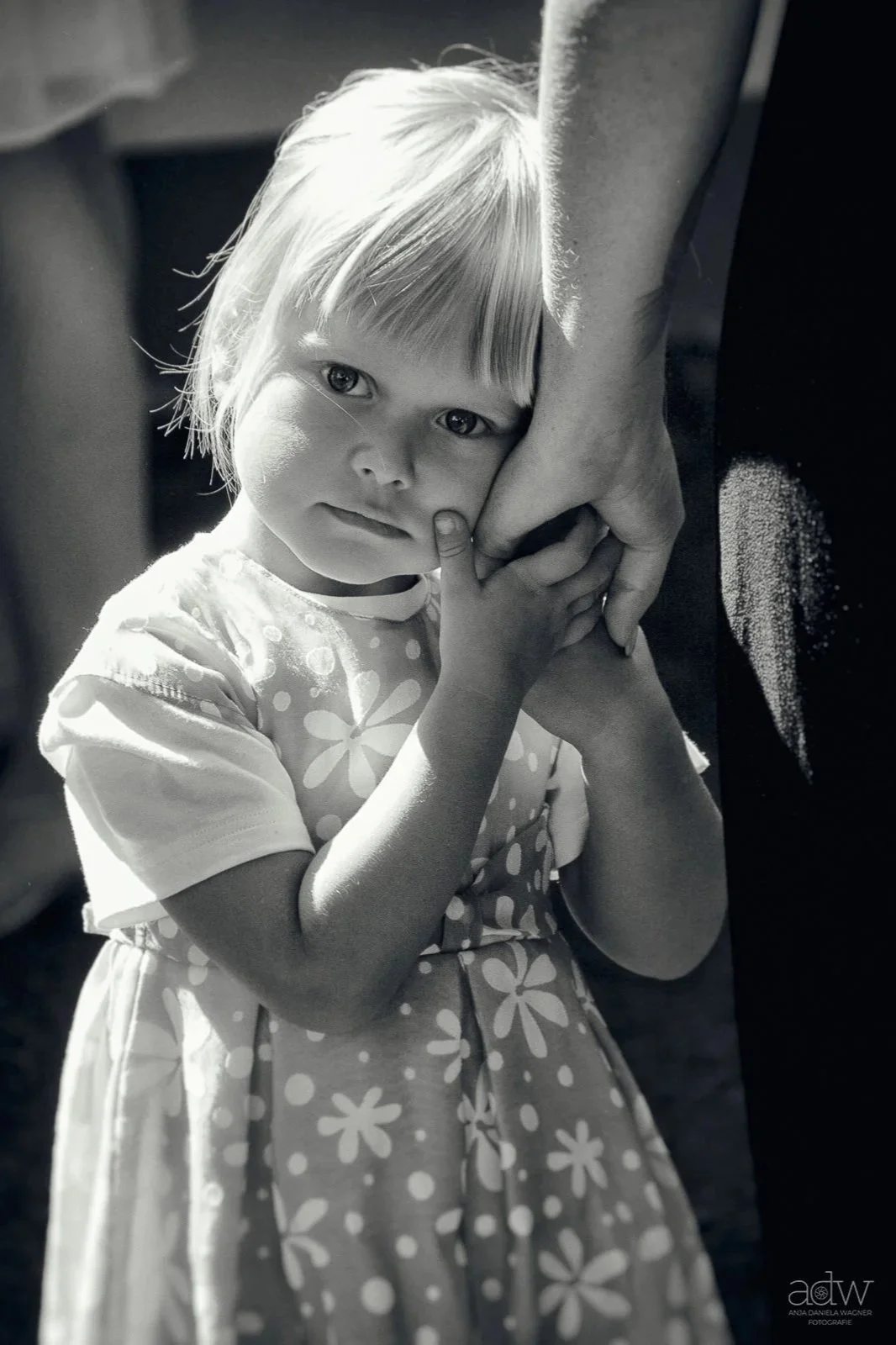 A young girl with light hair wearing a floral dress is holding hands with an adult, and she is touching her face with her finger, looking at the camera with a gentle expression in a black and white photograph.