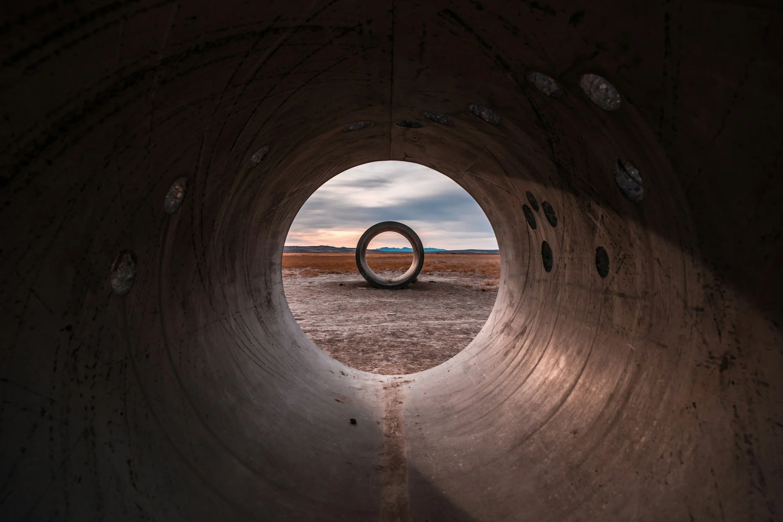 Looking through a large metal pipe at an open landscape with mountains in the distance, cloudy sky, and dry grass