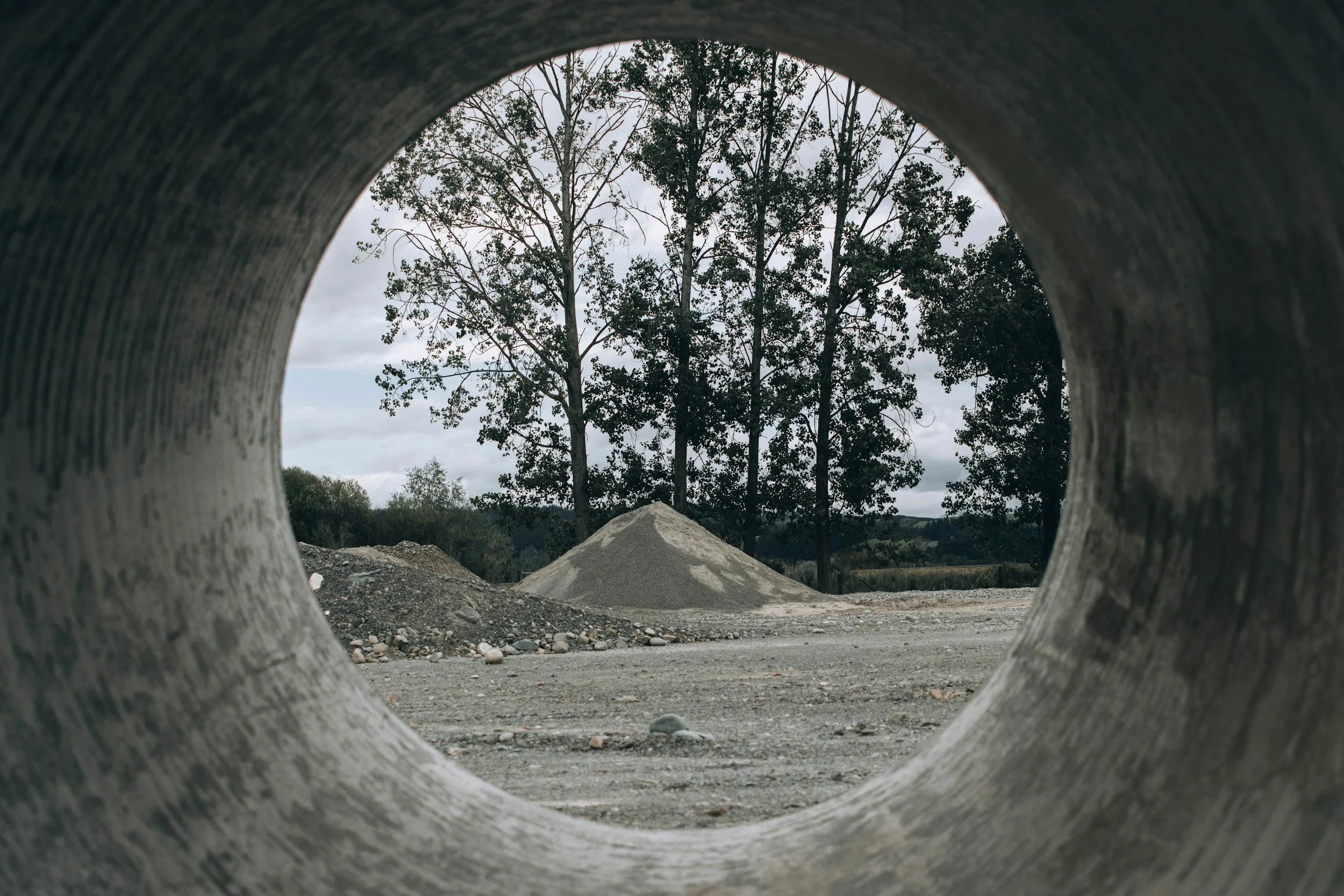 View of trees and dirt mounds seen through a circular hole in a concrete structure.