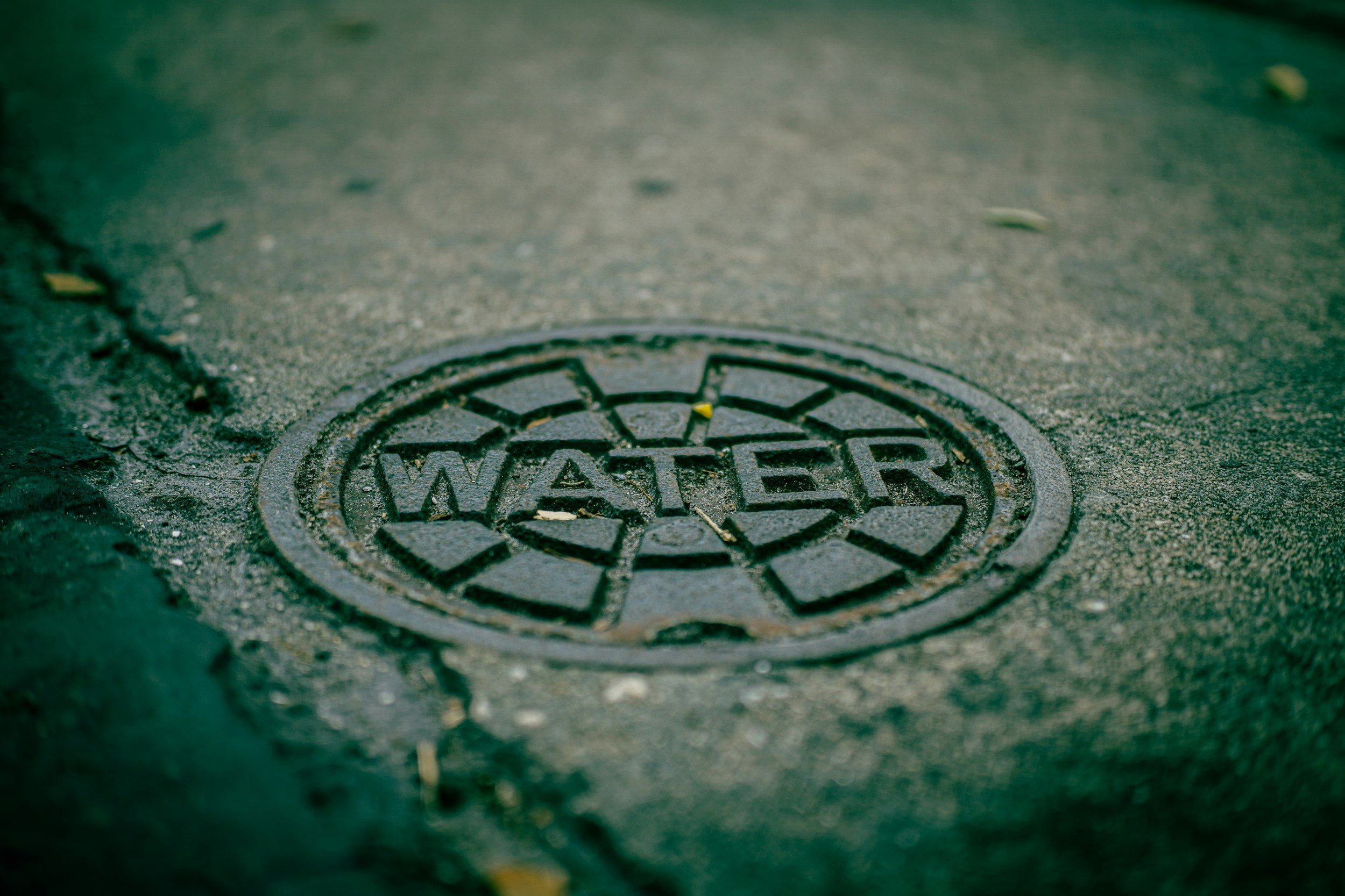 A circular water drain cover embedded in a weathered asphalt street surface with some small cracks and scattered debris.