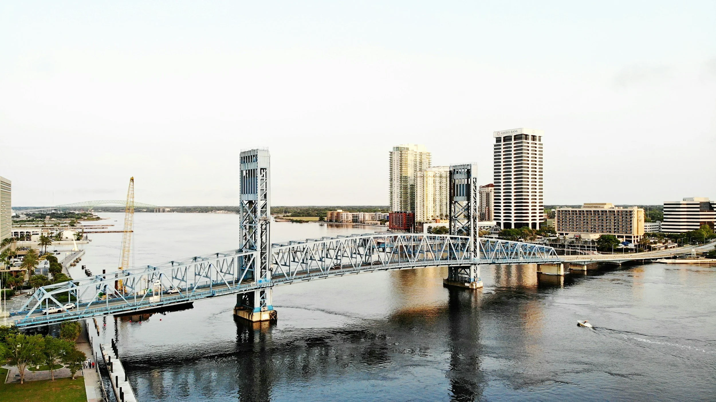 City skyline with tall buildings, a bridge spanning a river, and a boat on the water, with a clear sky overhead.