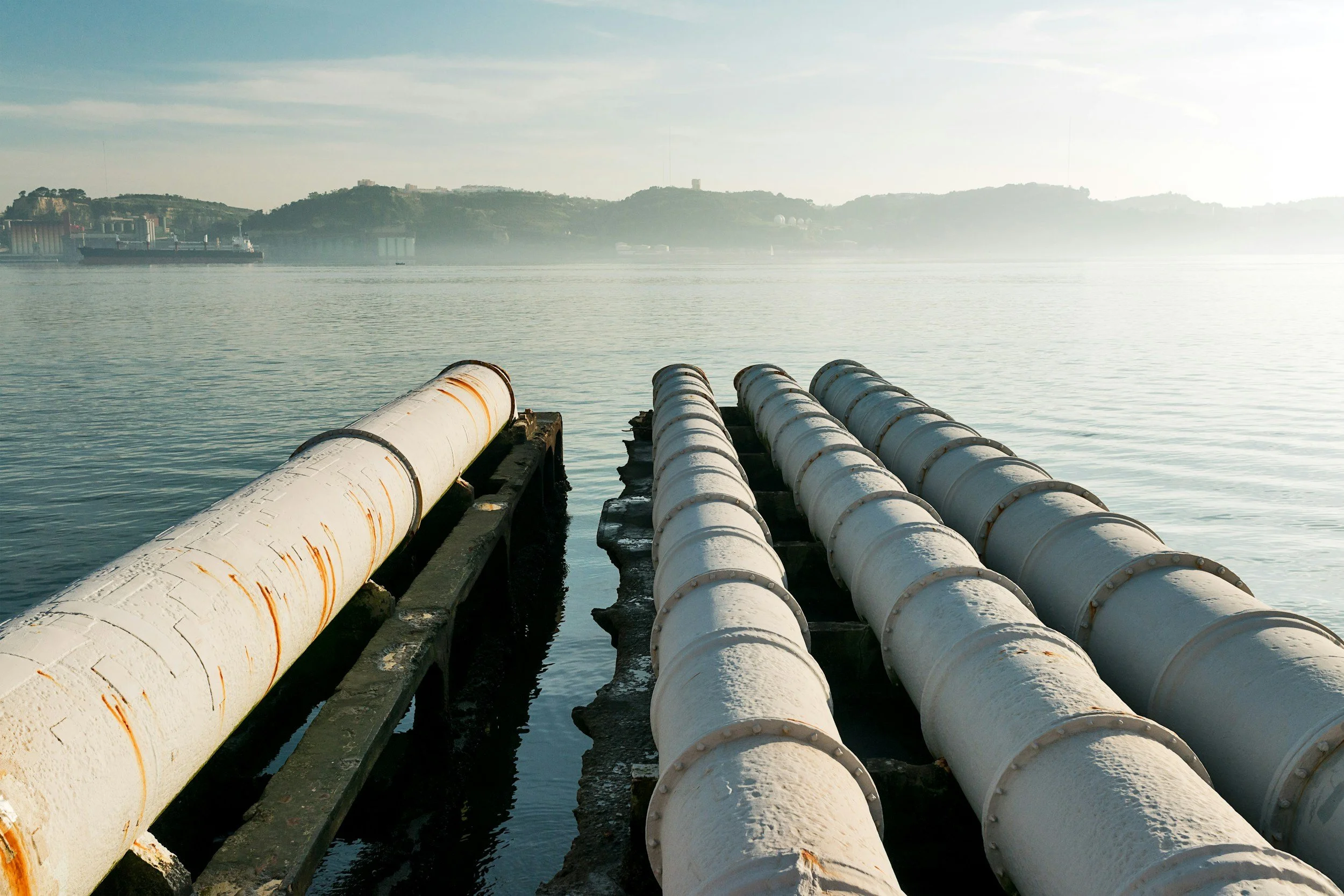 View of large white and rusted pipes on a structure extending into calm water with hazy hills and boats in the background.