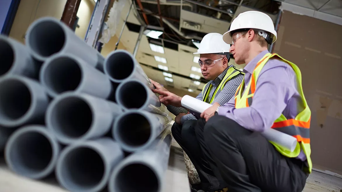 Two construction workers wearing safety helmets and vests examining pipes and blueprints inside a building under construction.