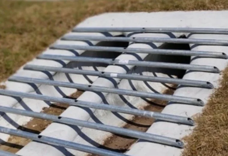 Drainage grate covering a tunnel with concrete walls and metal bars, installed on a patch of grass.
