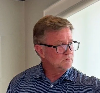 A man with glasses and light brown hair looking to the side, wearing a blue striped shirt, standing indoors near a window.