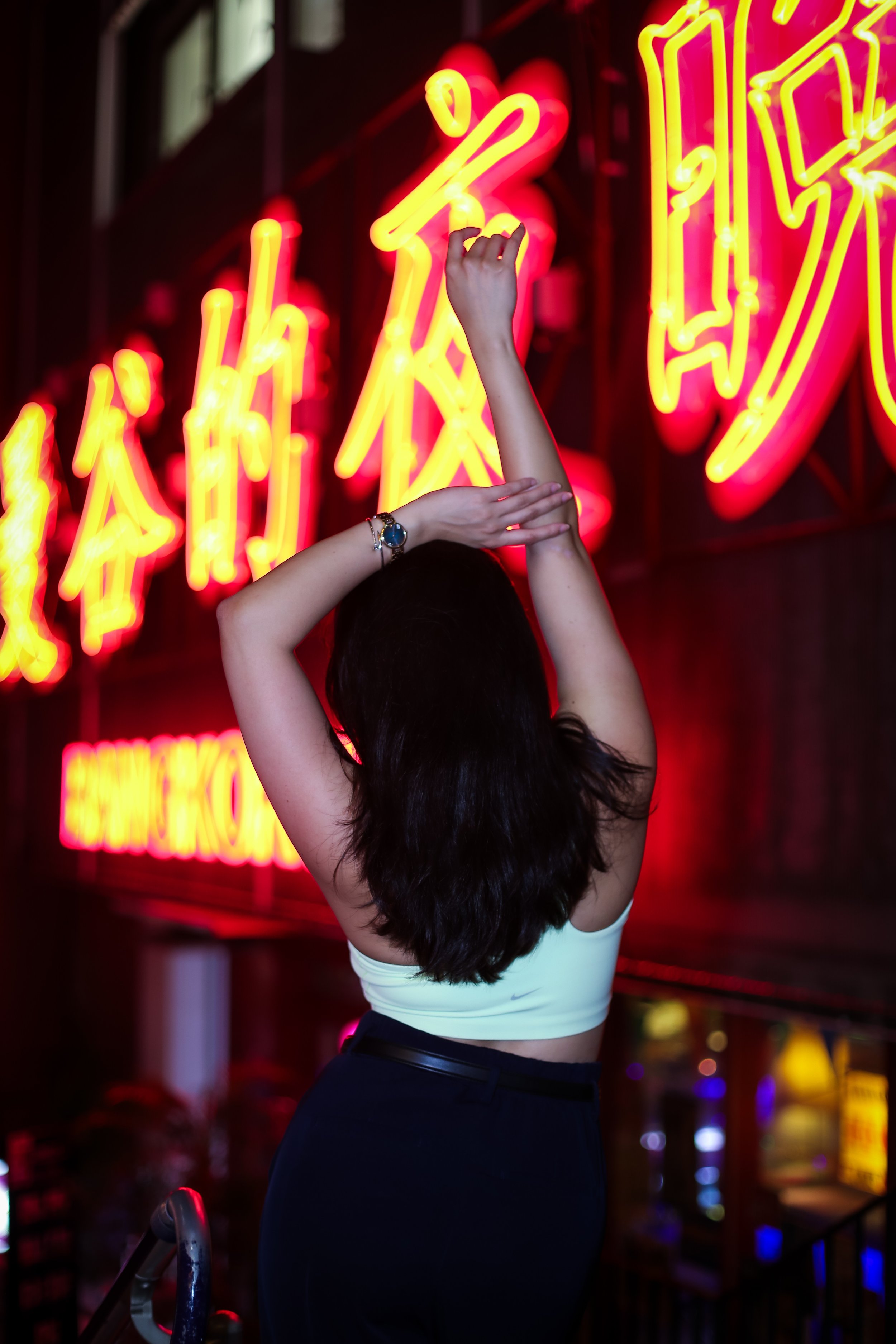 A woman with dark hair, wearing a white sports bra and black pants, is standing in front of a bright neon sign with yellow and red Chinese characters, reaching up with one arm towards the sign in a dimly lit environment.