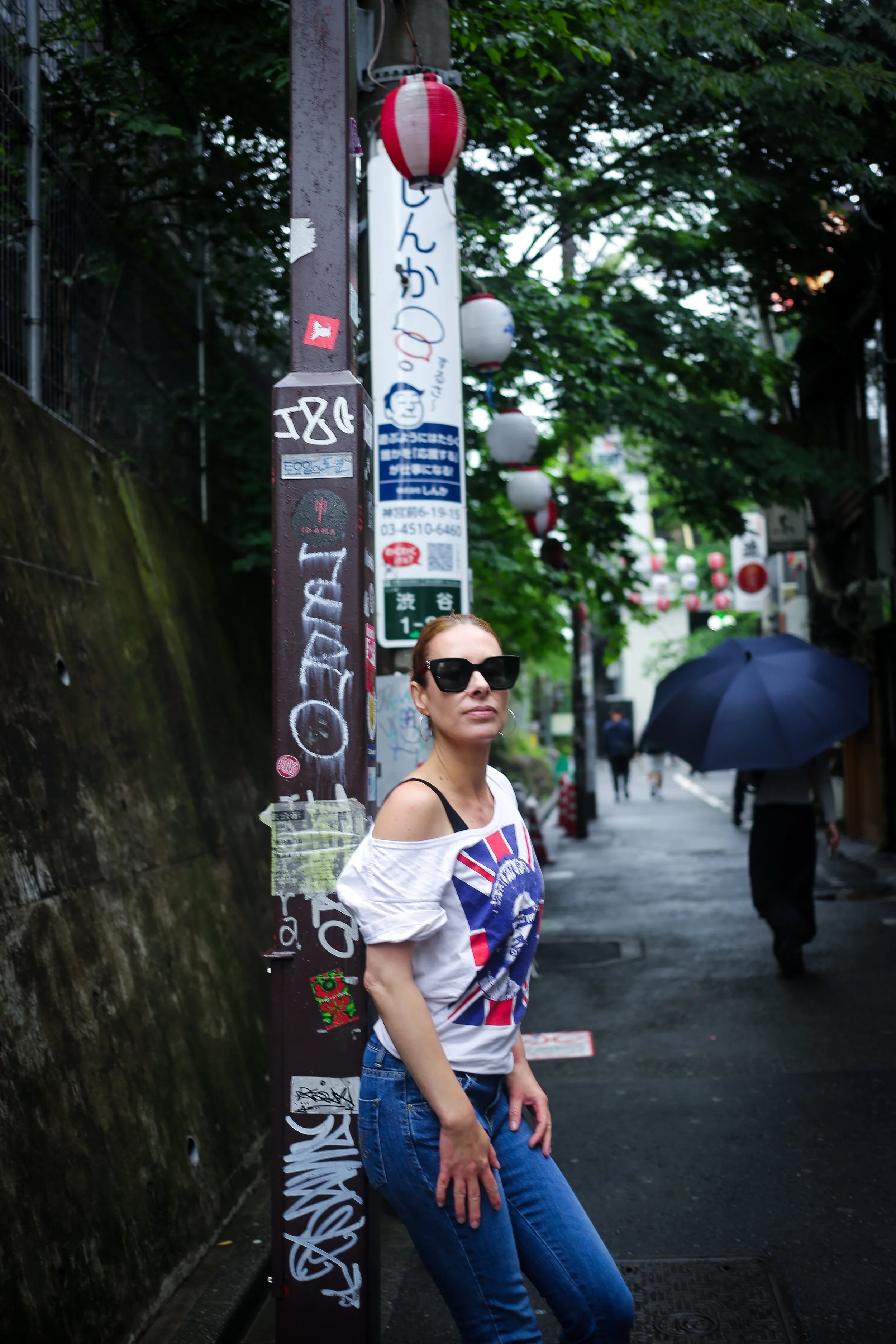 A woman wearing sunglasses, a t-shirt with a Union Jack design, and blue jeans standing on a street corner with a pole covered in stickers and graffiti, holding her leg and with umbrellas and pedestrians in the background.