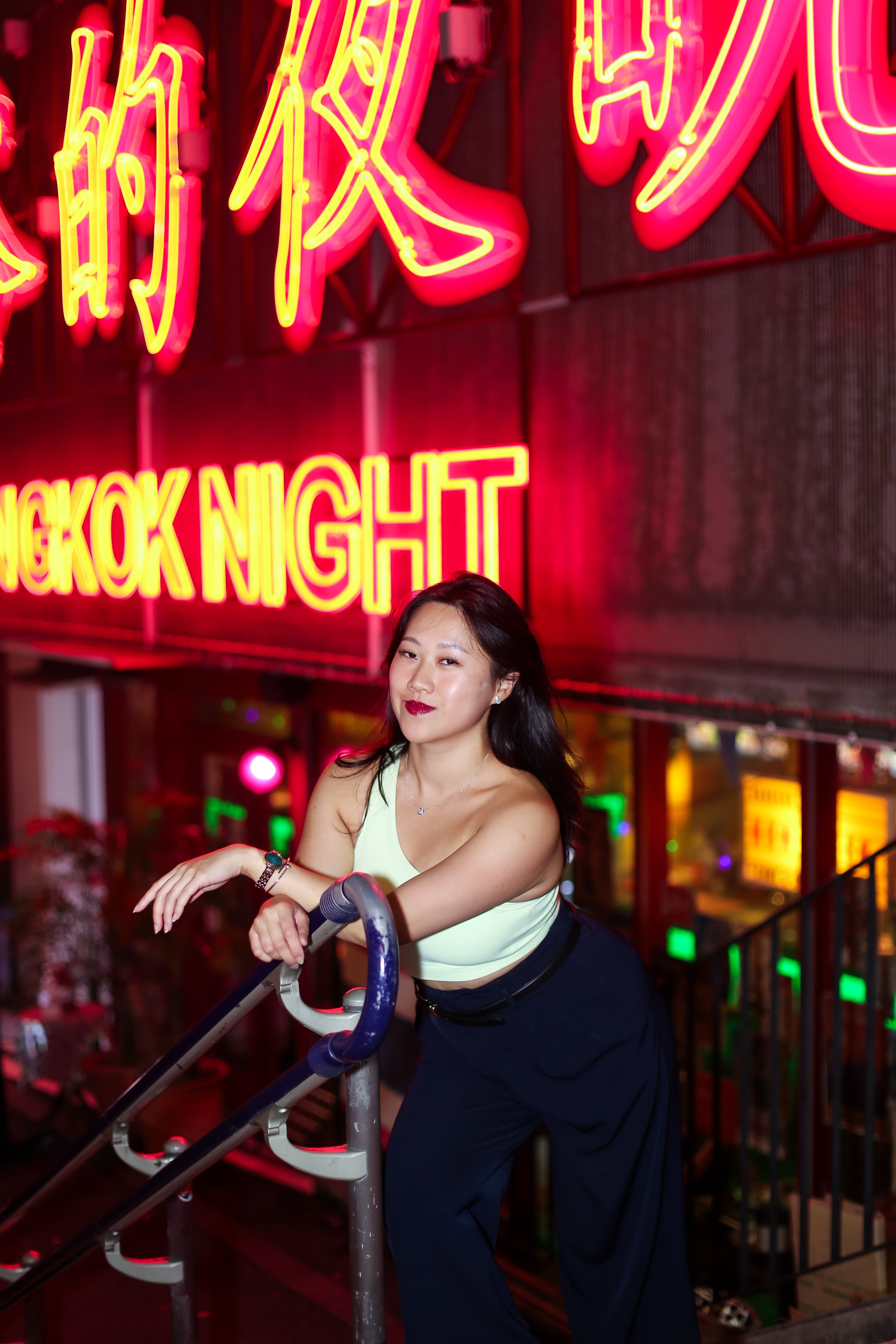 A woman with long dark hair smiling while leaning on a scooter in front of neon signs that read 'Bangkok Night' at night.