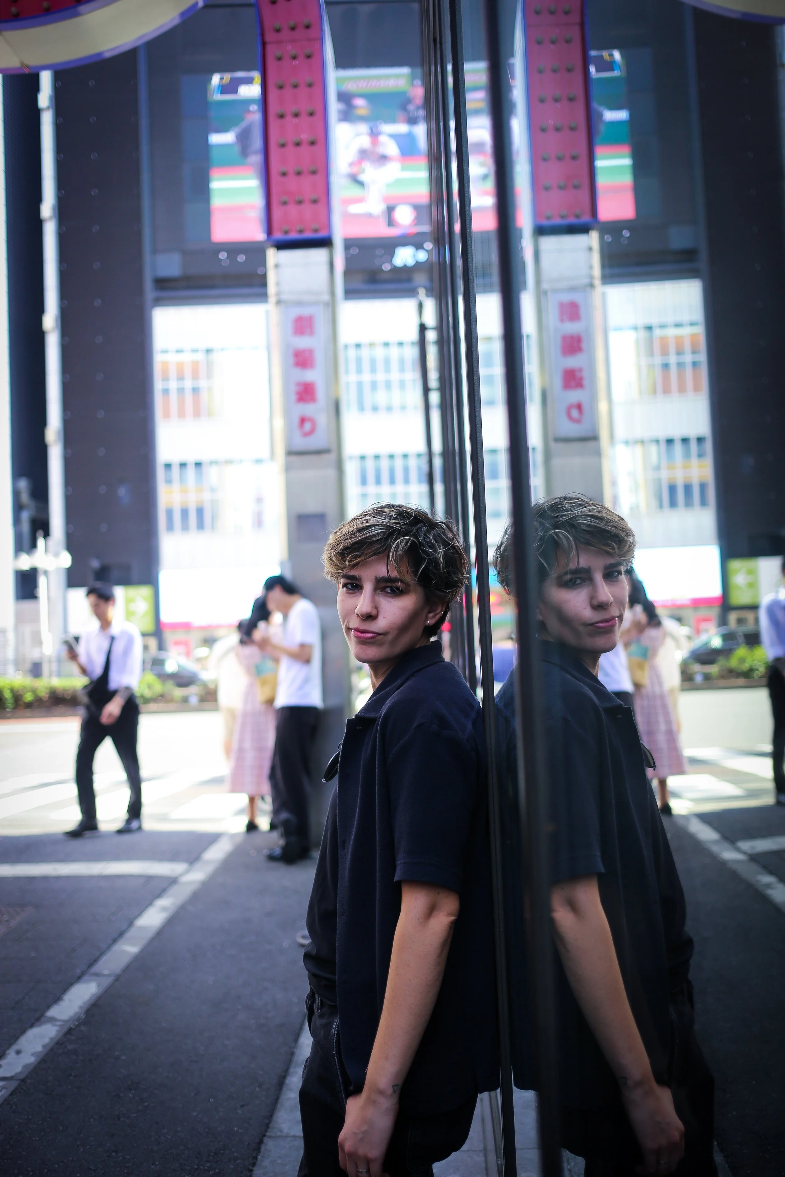 A young woman with short hair and a dark shirt standing in front of a mirror at an urban outdoor location. The woman's reflection is visible in the mirror. Background shows pedestrians, buildings, and digital screens, indicating a city setting.