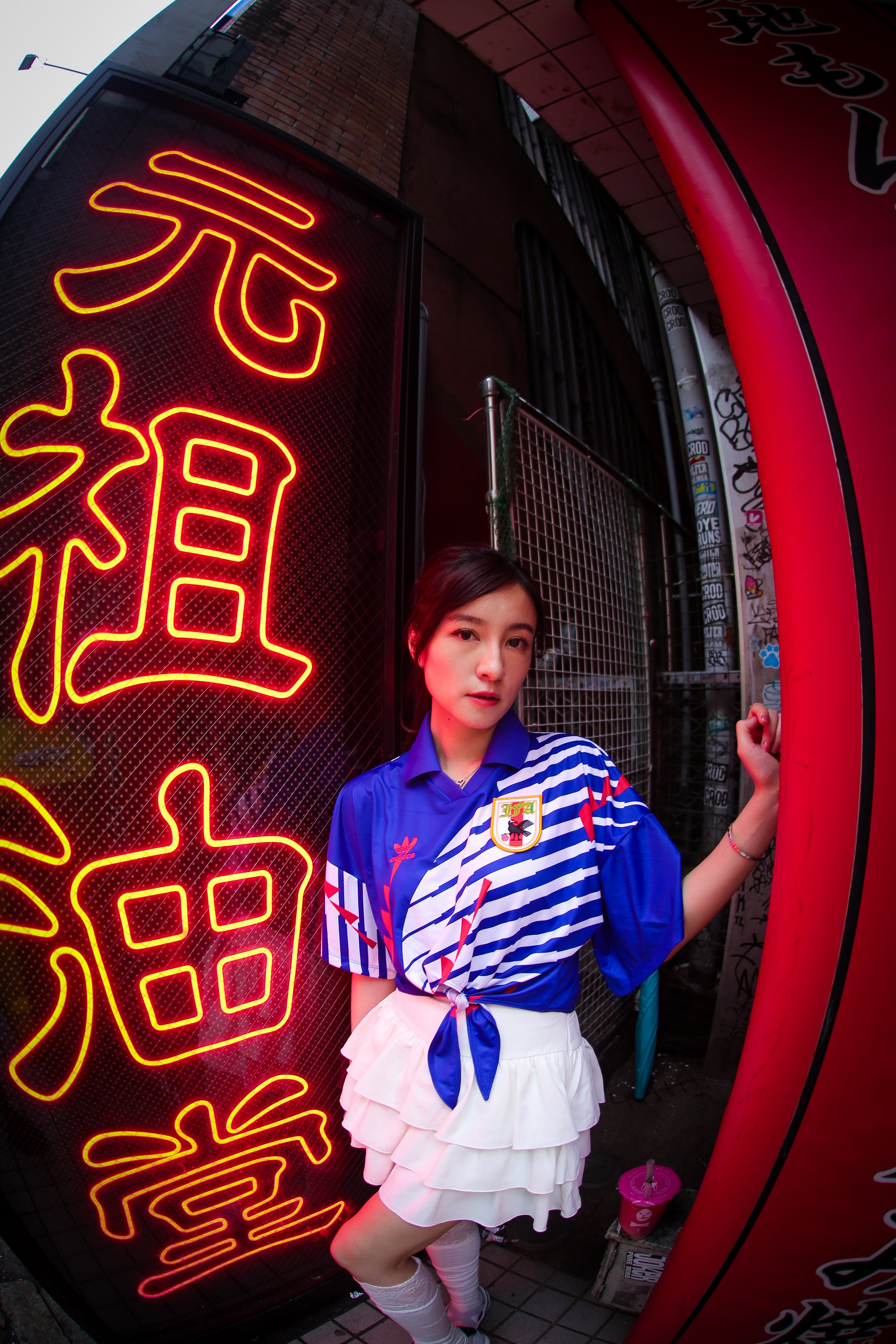 Young woman standing near a neon sign with Chinese characters, dressed in a blue sports shirt with a crest, white ruffled skirt, and thigh-high socks, holding a pink cup in her hand.