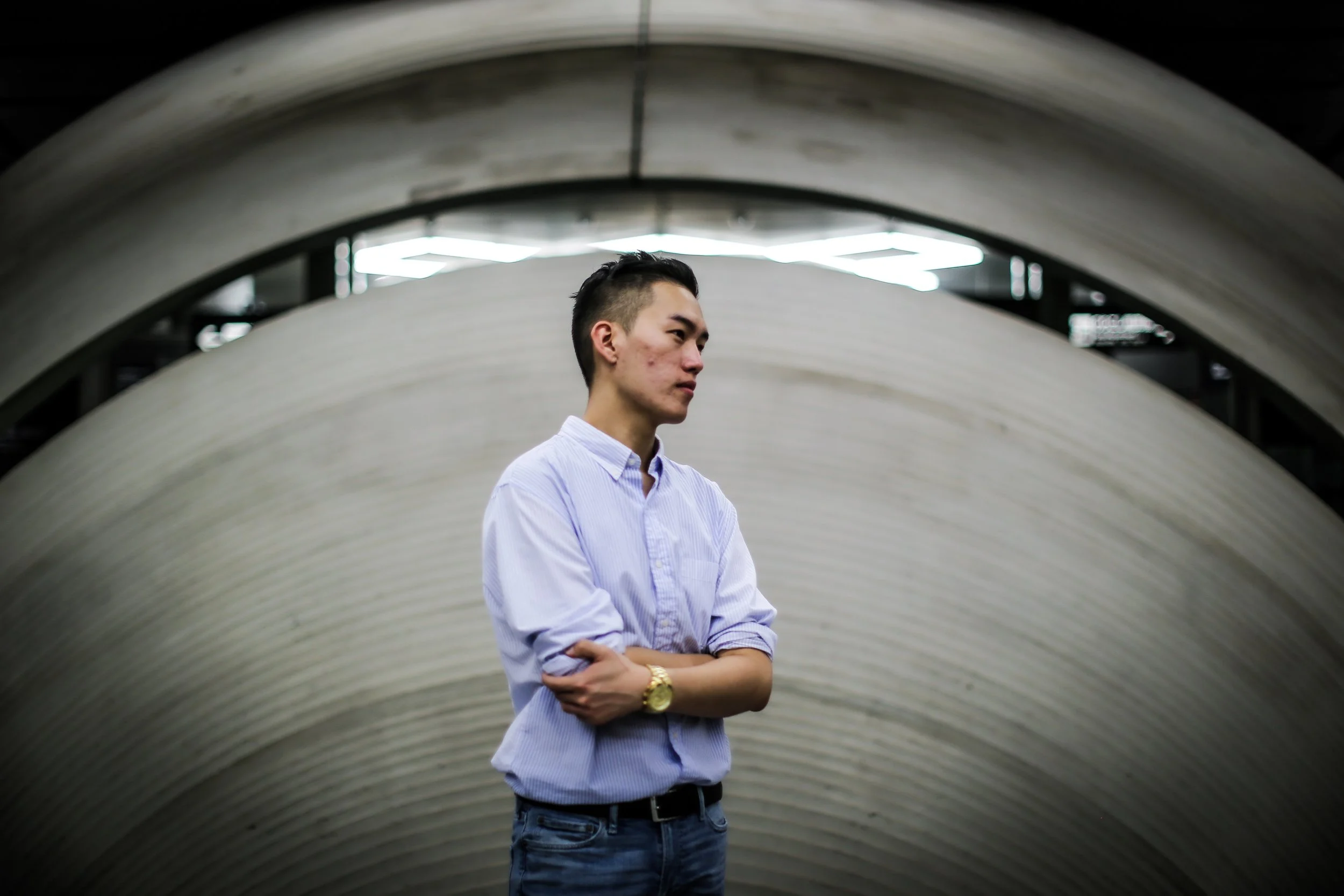 A young man in a light blue striped shirt with rolled-up sleeves and a gold wristwatch stands with his arms crossed in front of an abstract, curved architectural background with geometric lighting.