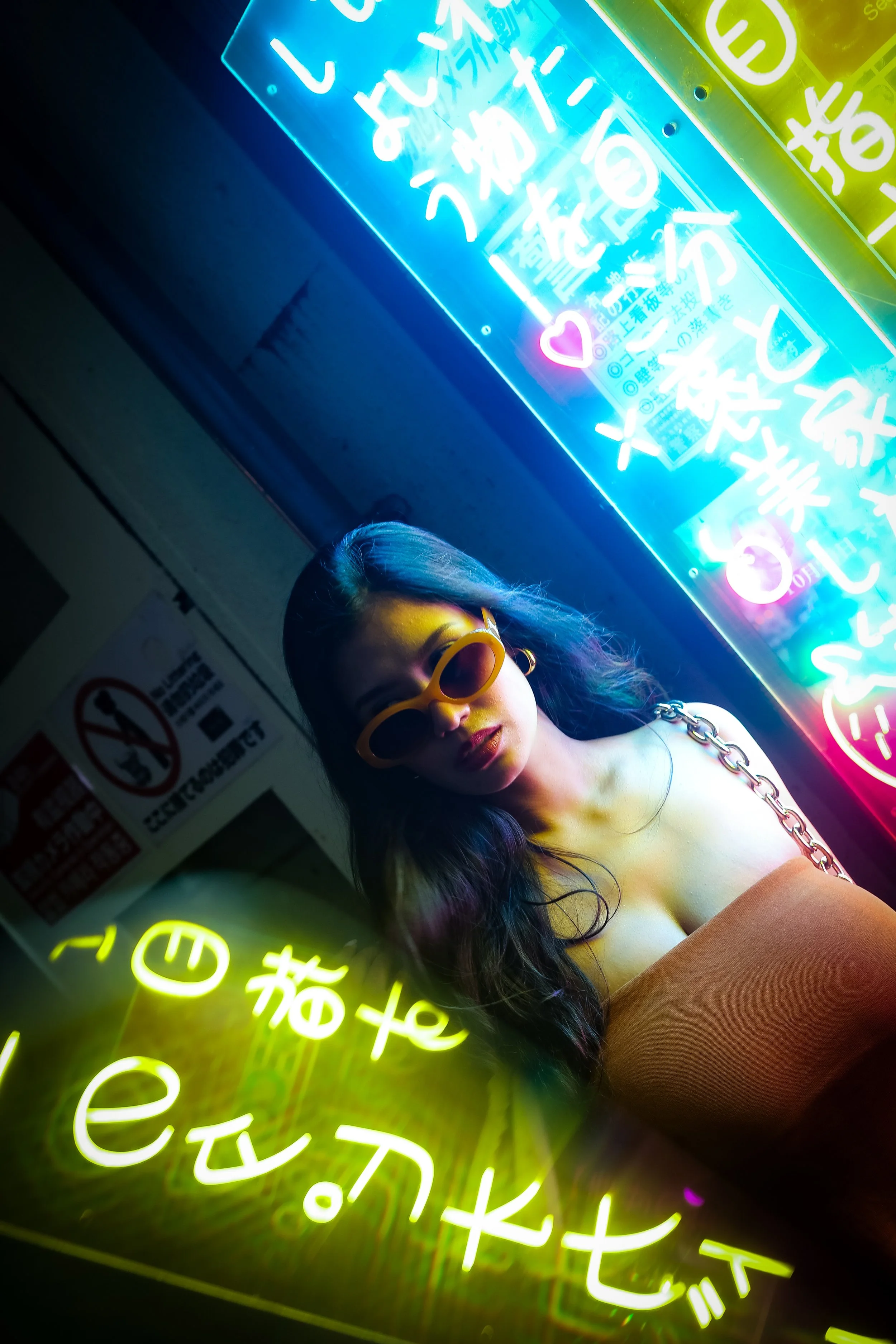 A woman with long dark hair wearing orange sunglasses and a chain strap top, standing in front of neon signs with Japanese characters and symbols, illuminated with bright blue, pink, and yellow colors.