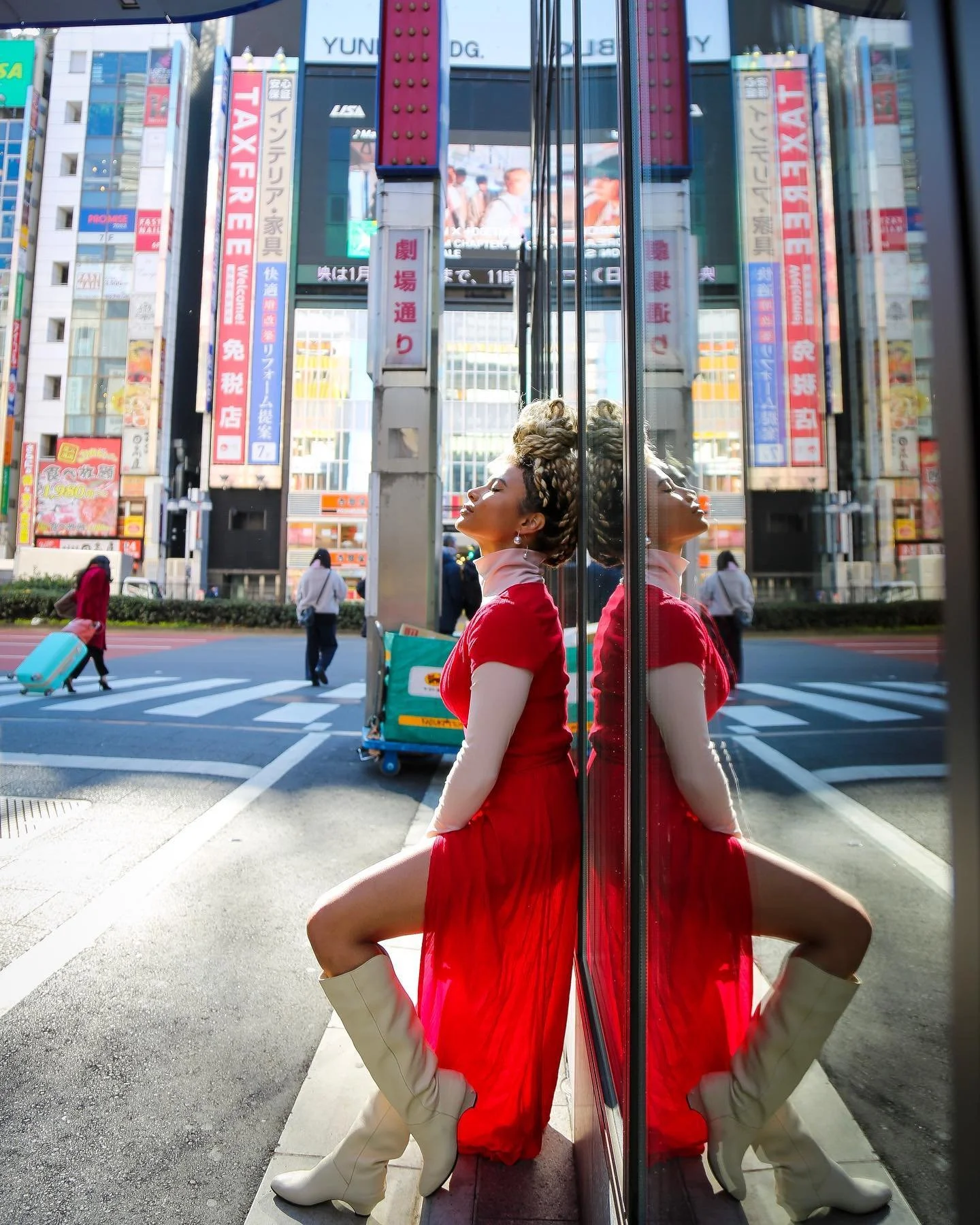 A woman in a red dress and beige boots is sitting with her back against a reflective glass wall, with her reflection visible. She is outdoors in an urban area with signs and people passing by, and the scene is illuminated by daylight.