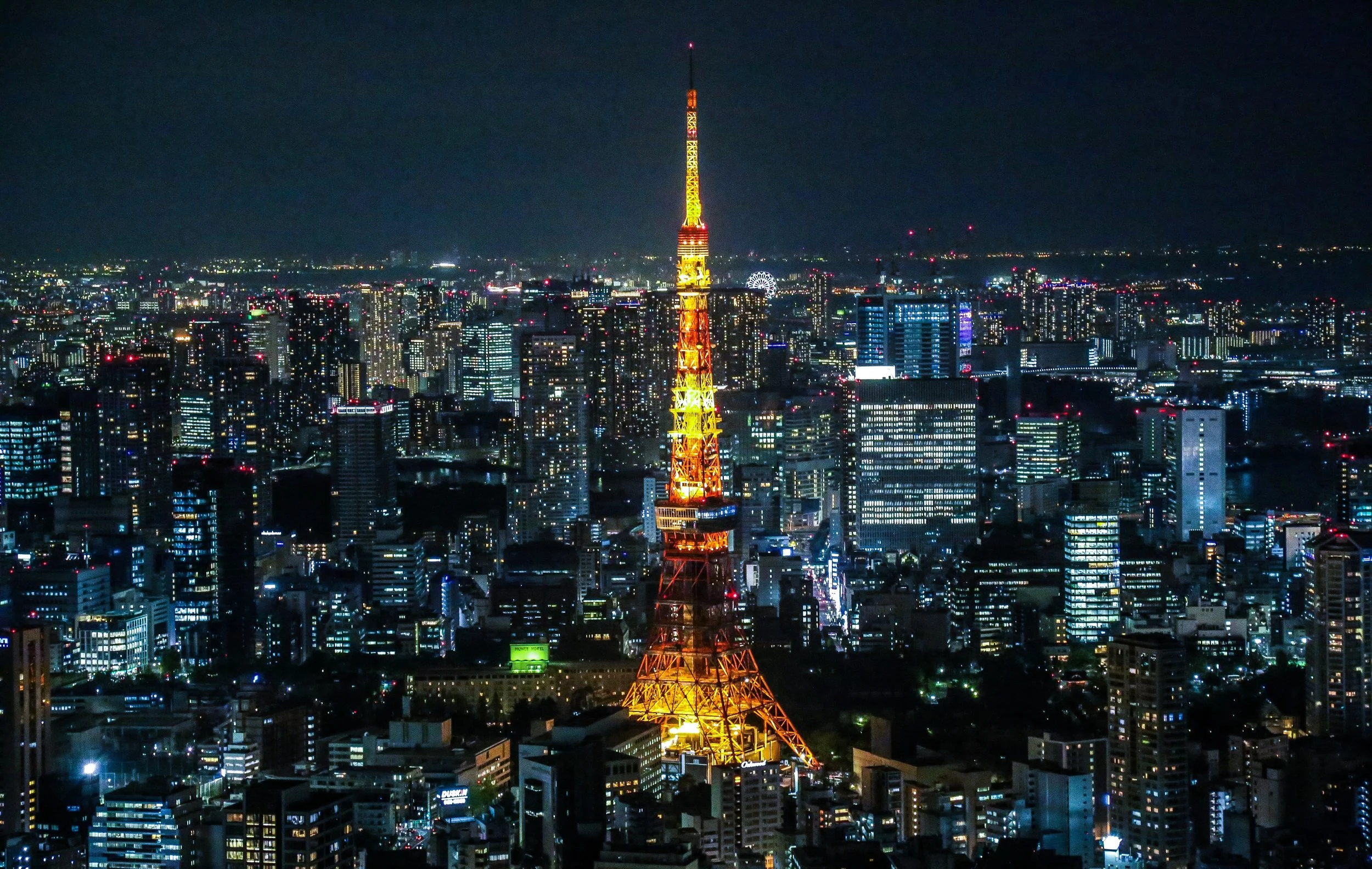Night view of a city skyline with a brightly lit, orange-golden communication tower in the foreground. City buildings and a ferris wheel are visible in the background.