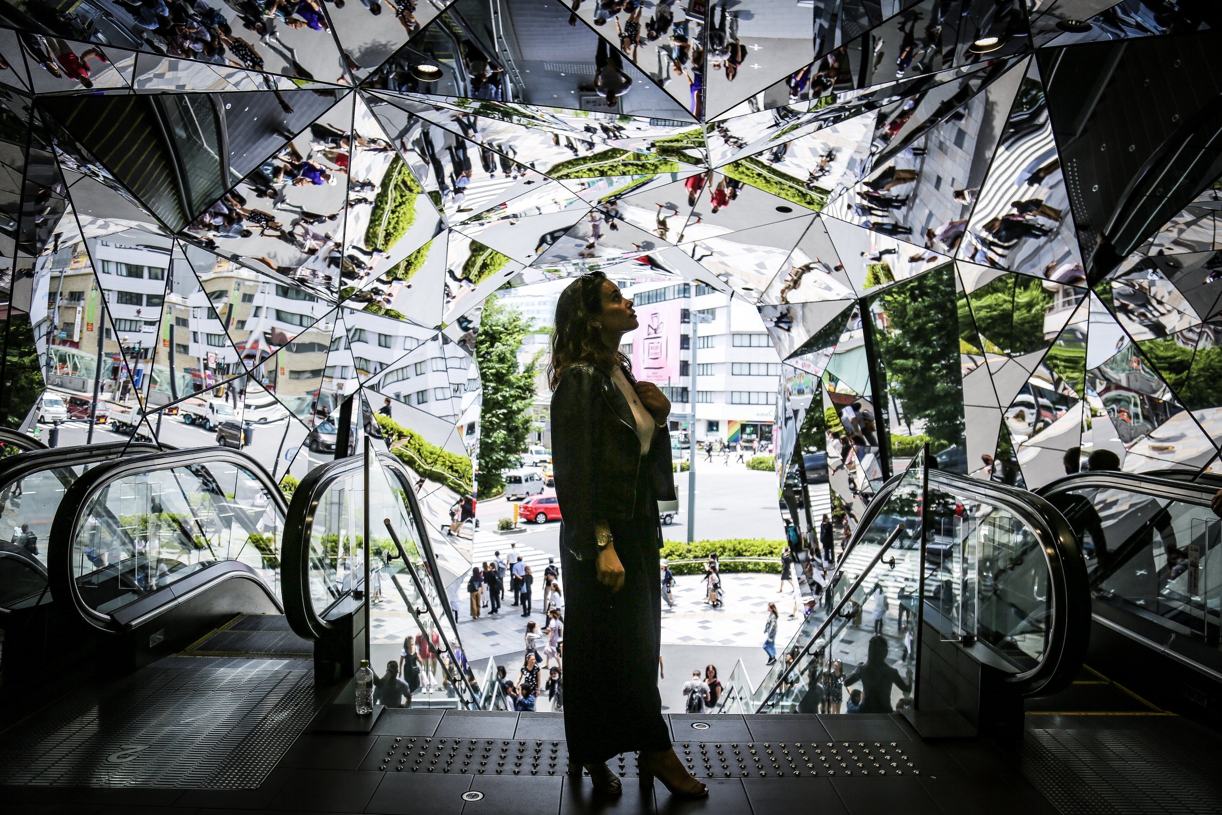 A woman stands in an artistic mirrored installation, looking out at a bustling city street filled with pedestrians, cars, and buildings.