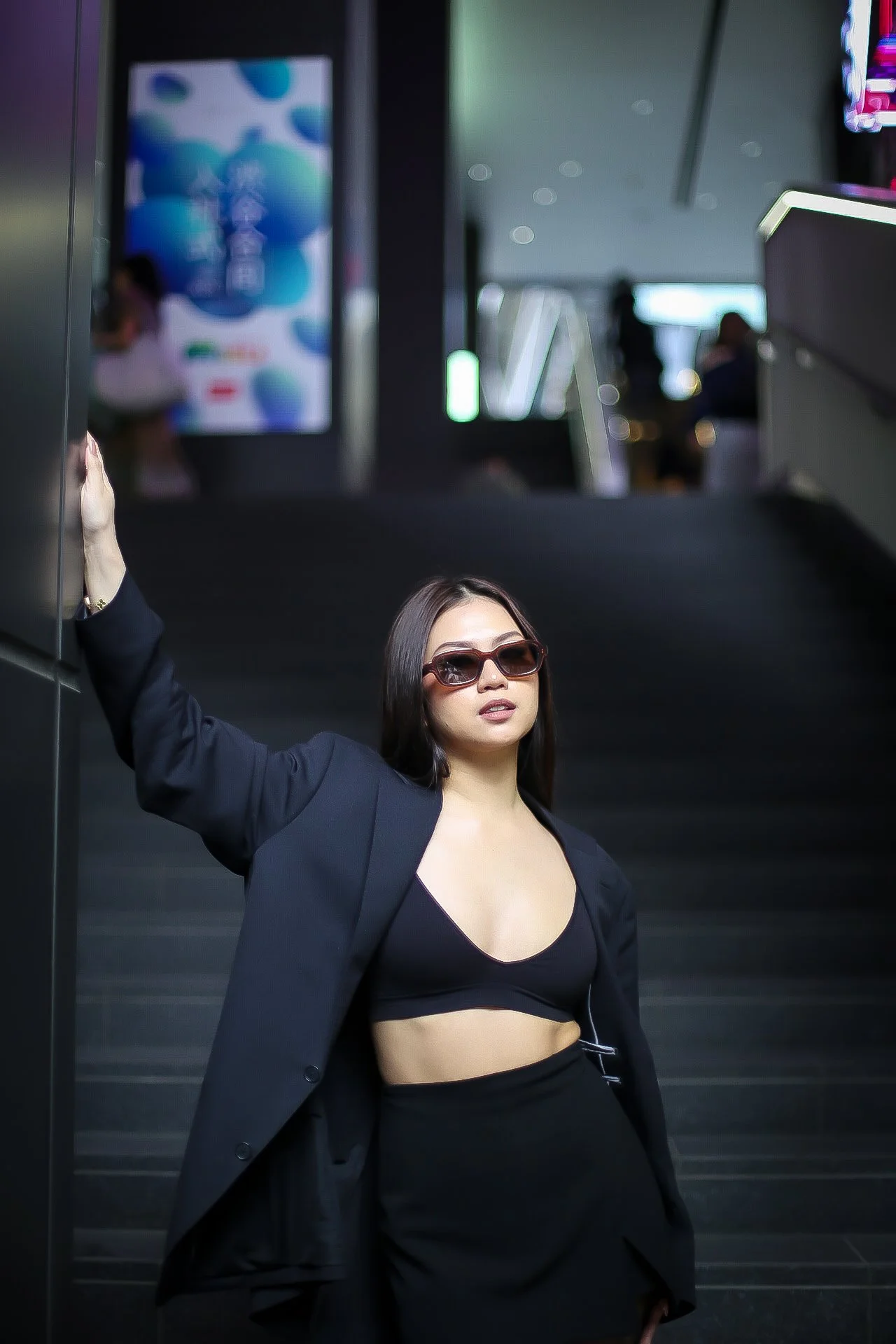 A woman in a black blazer and crop top with sunglasses posing with her left arm raised inside a modern building, possibly an airport or shopping mall.