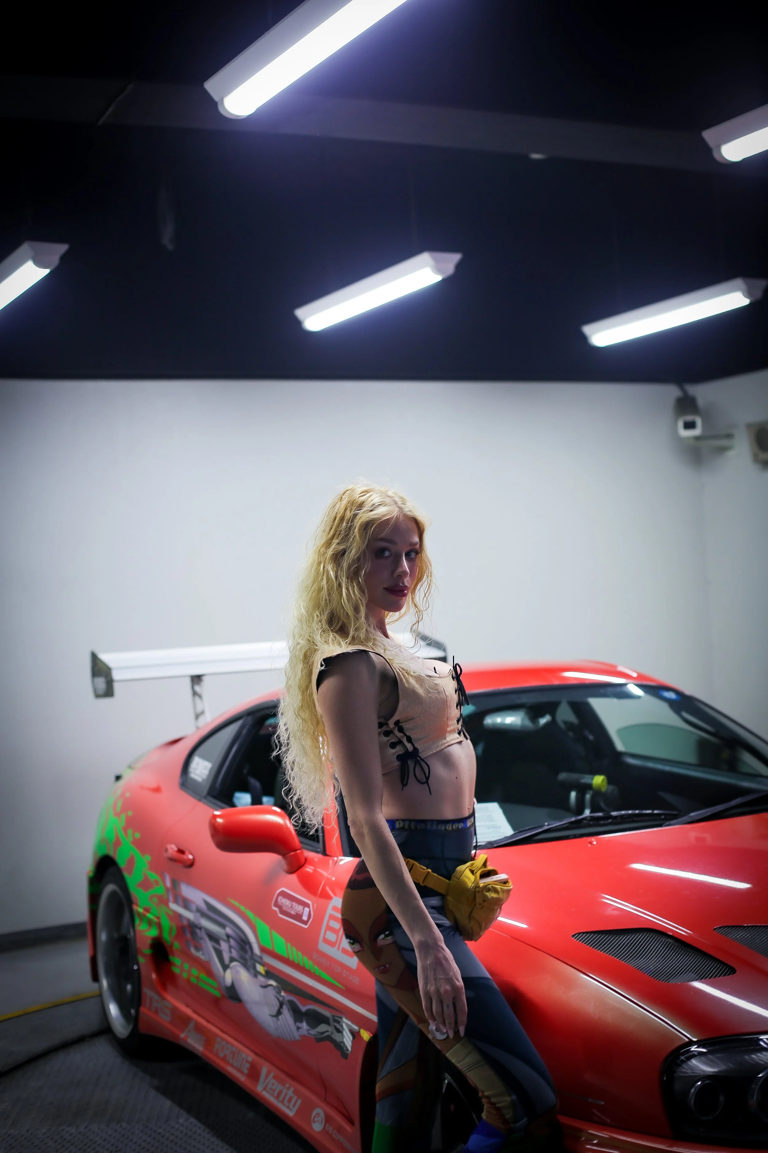 A woman with long curly blonde hair standing next to a red race car inside a garage with white walls and fluorescent lights above.