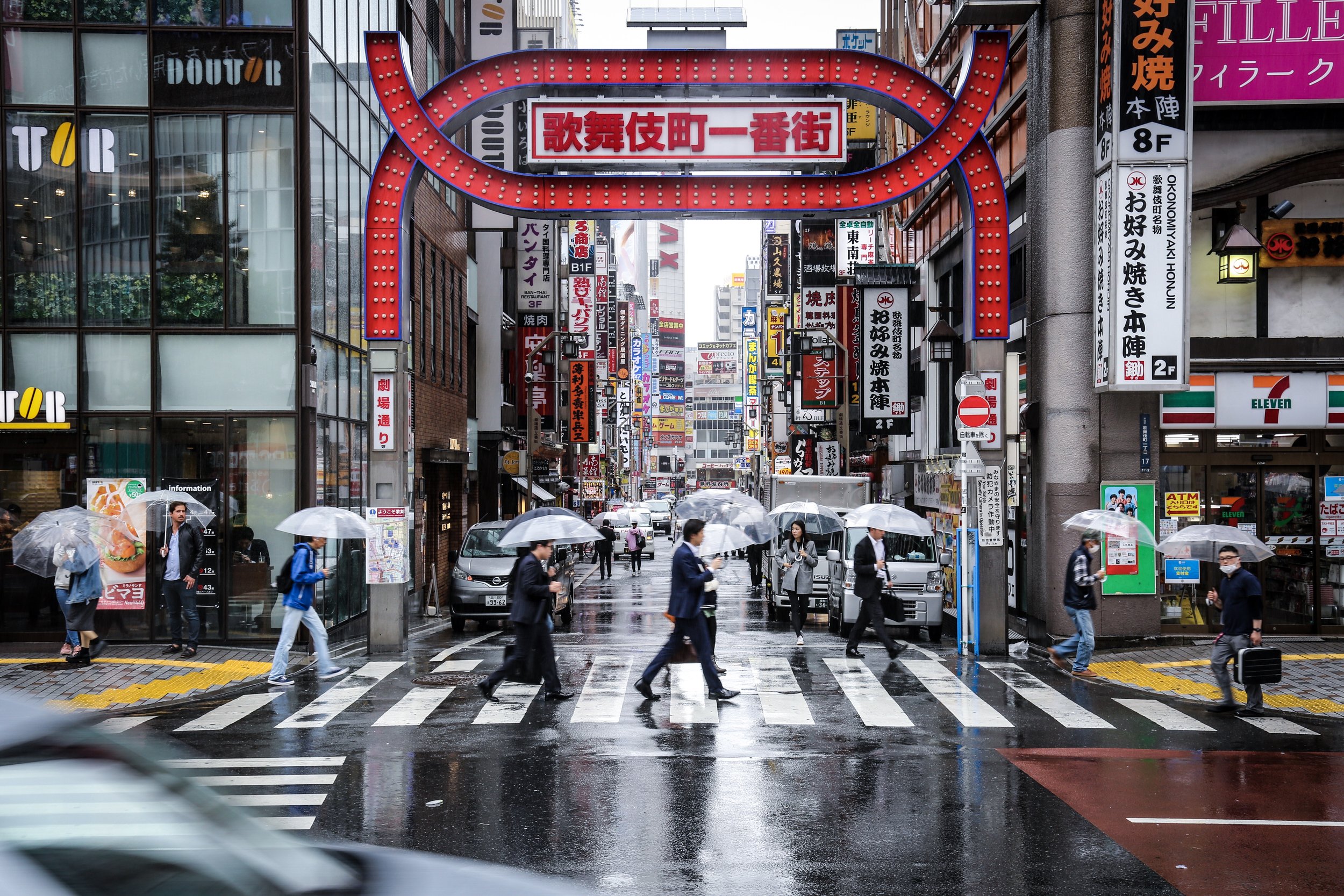 Street scene with pedestrians carrying umbrellas on a rainy day in an urban area in Japan, featuring numerous signs and advertisements in Japanese.