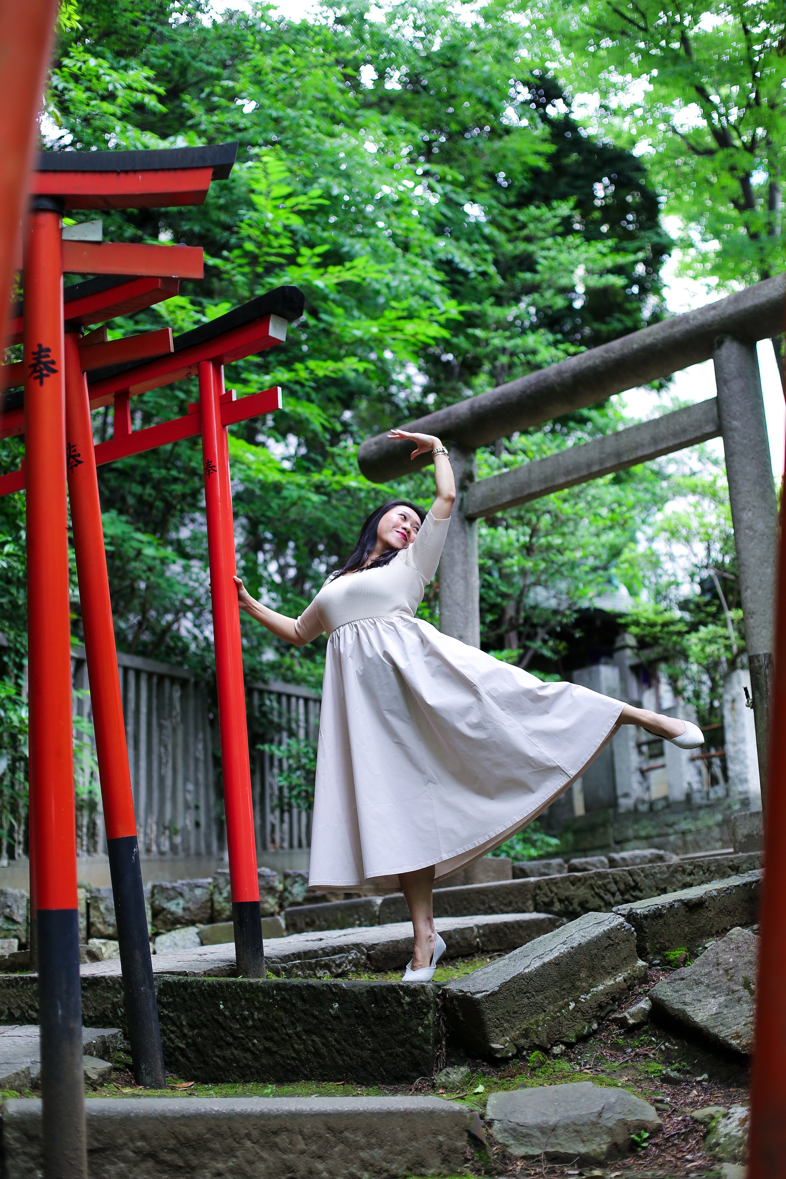 A woman in a beige dress and high heels doing a ballet pose on stone steps near red and gray shrine torii gates in a lush green outdoor setting.