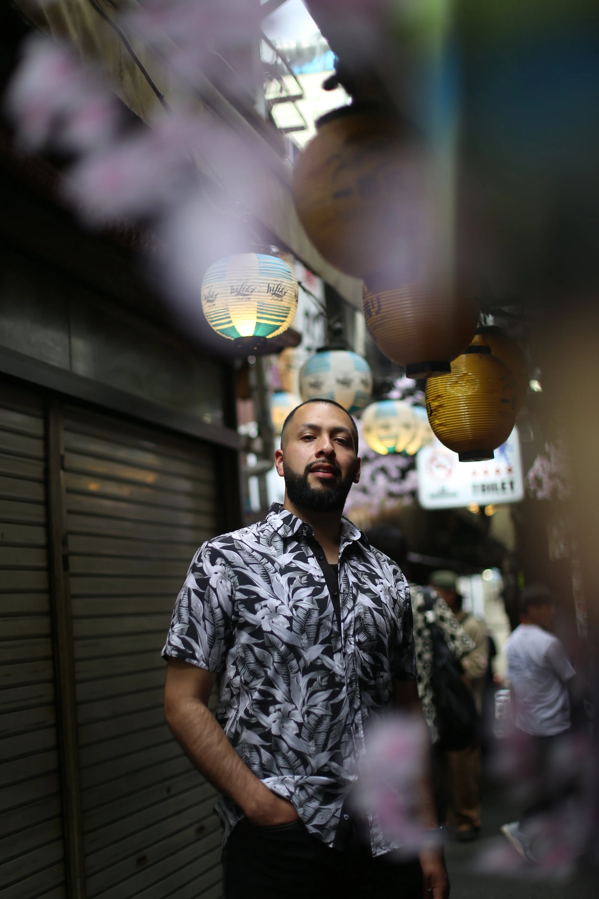Man with a beard wearing a white and black floral patterned shirt standing outside under hanging lanterns during dusk or evening.