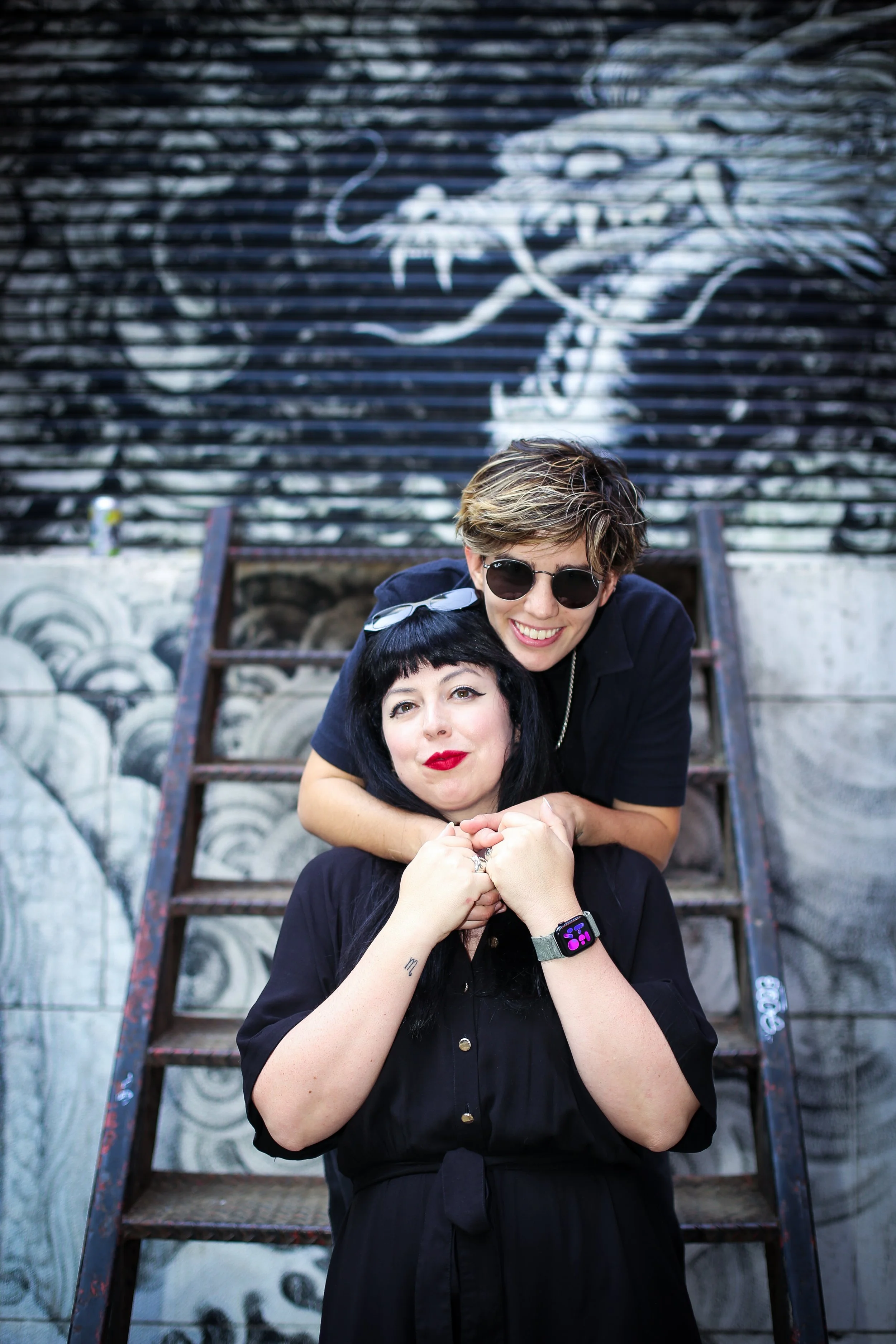 Two women smiling, one with short blonde hair and sunglasses, the other with long dark hair and red lipstick, standing on graffiti-covered stairs with a black and white mural of a dragon in the background.