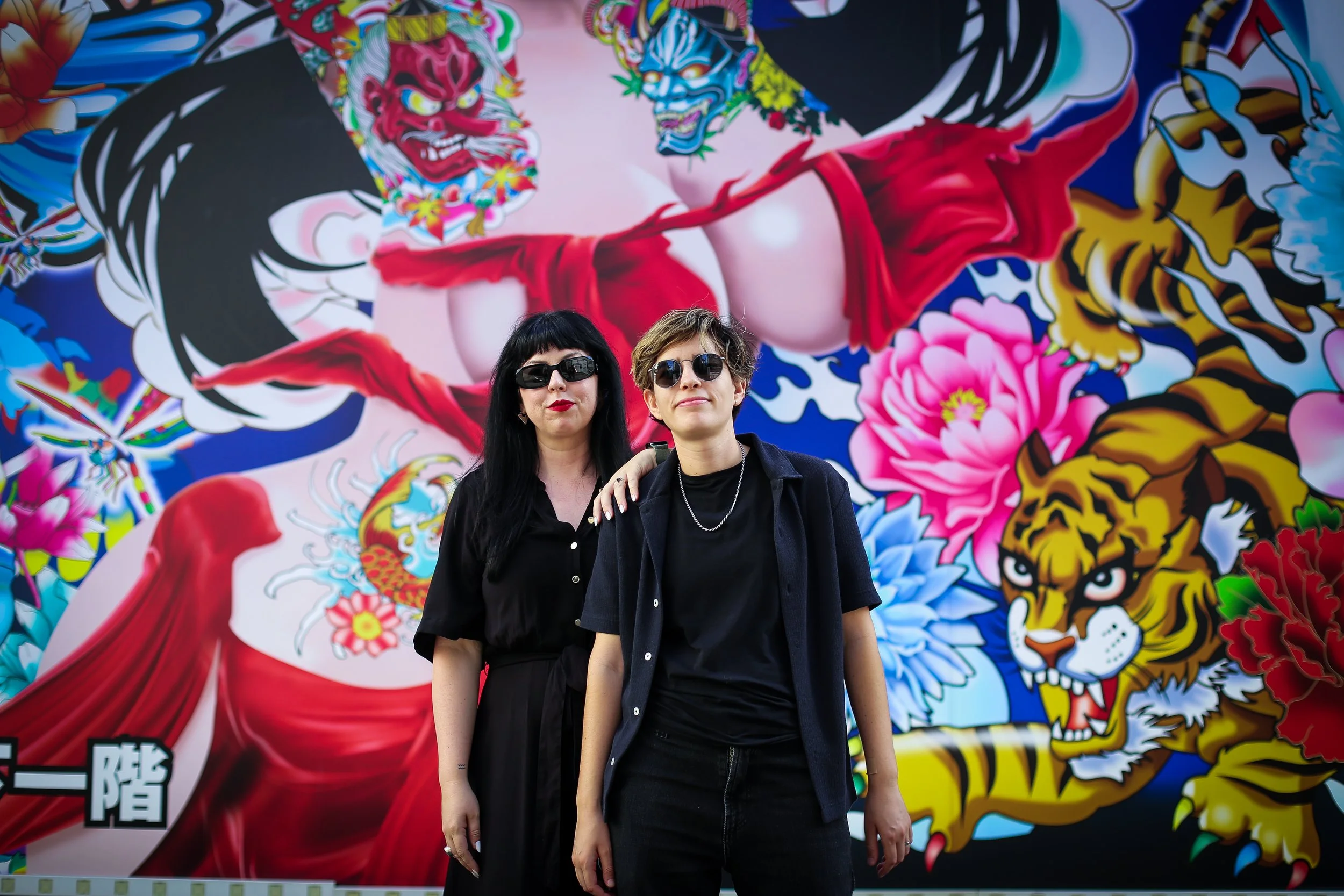 Two women standing in front of a large, colorful mural featuring a traditional Japanese tattoo style with a red demon, a tiger, and various floral and cloud elements. Both women are wearing black clothing and sunglasses.