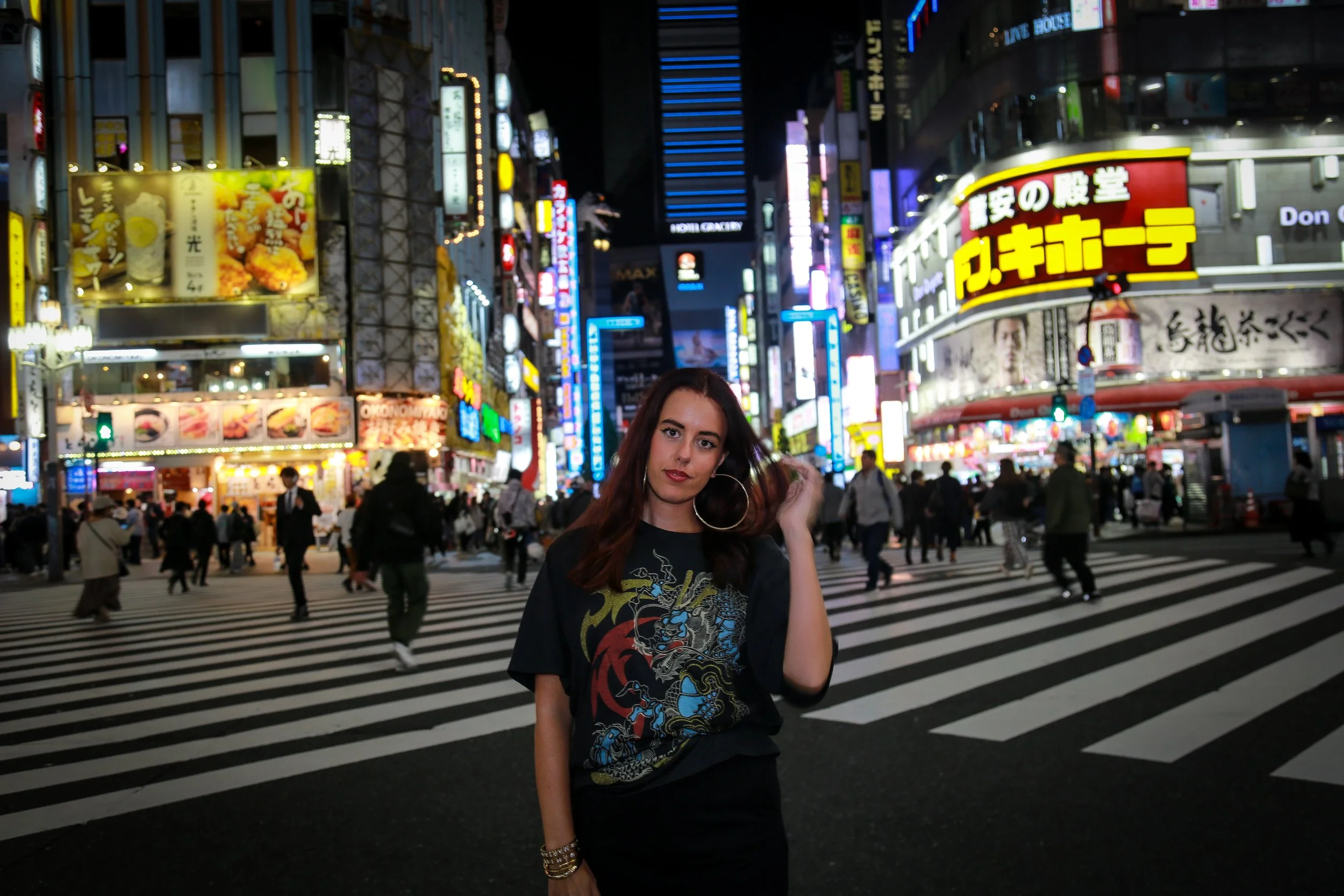 A woman with dark hair and large hoop earrings standing in front of a busy city intersection at night, with bright neon signs and advertisements illuminating the background.