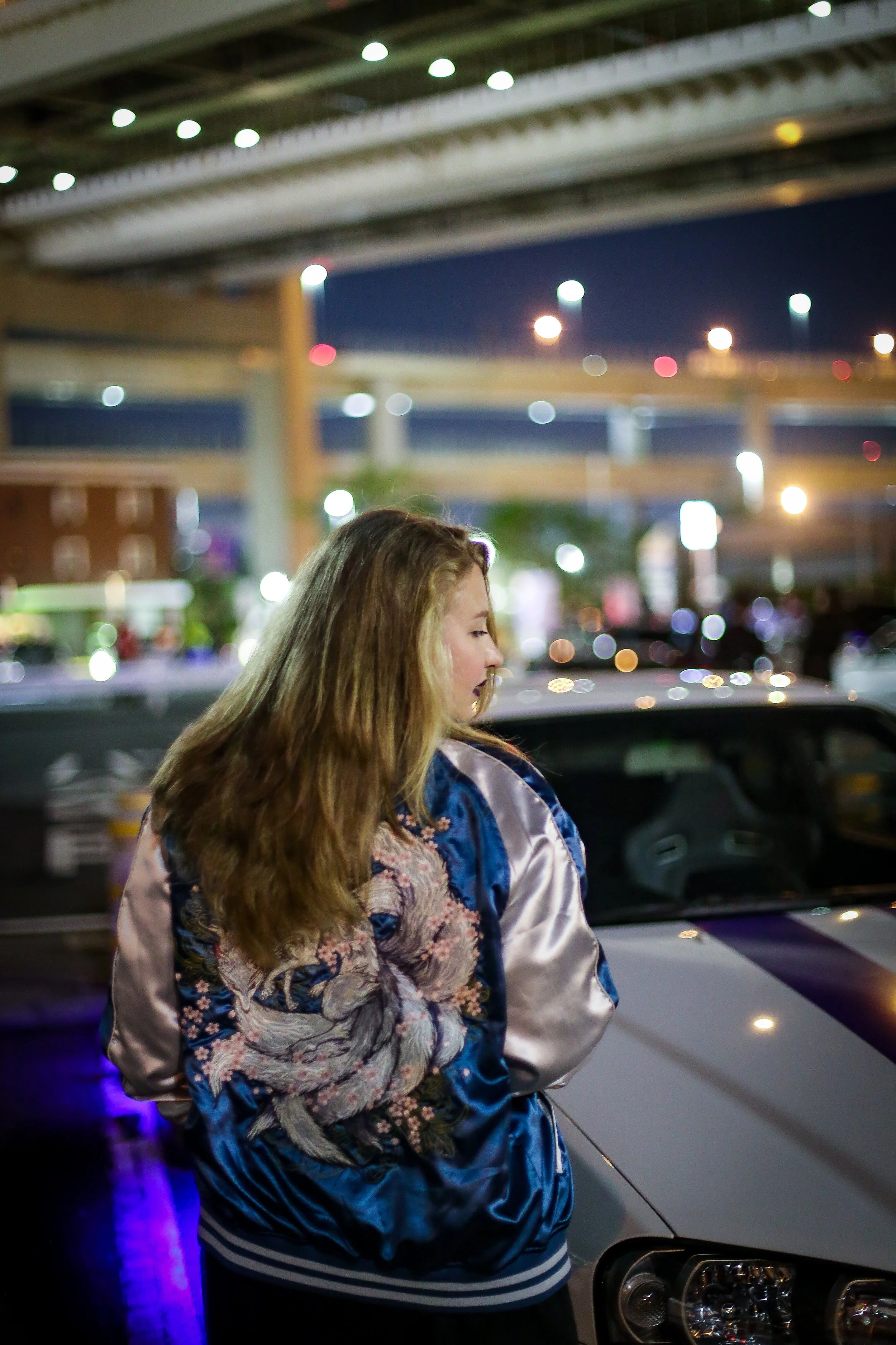 A woman with long wavy brown hair wearing a colorful floral and satin bomber jacket and striped shirt standing in front of a car at night with blurred city lights and an overpass in the background.