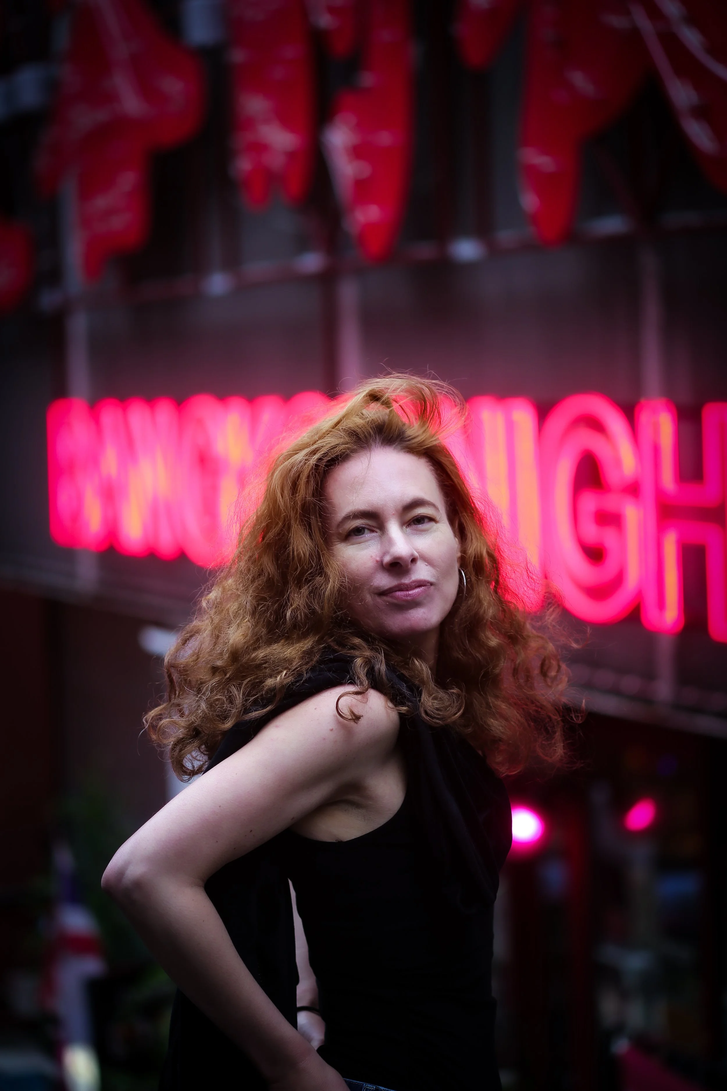 A woman with curly red hair standing outdoors at night in front of a neon pink sign that reads 'Dancing.' She is wearing a black sleeveless top and has a neutral expression.