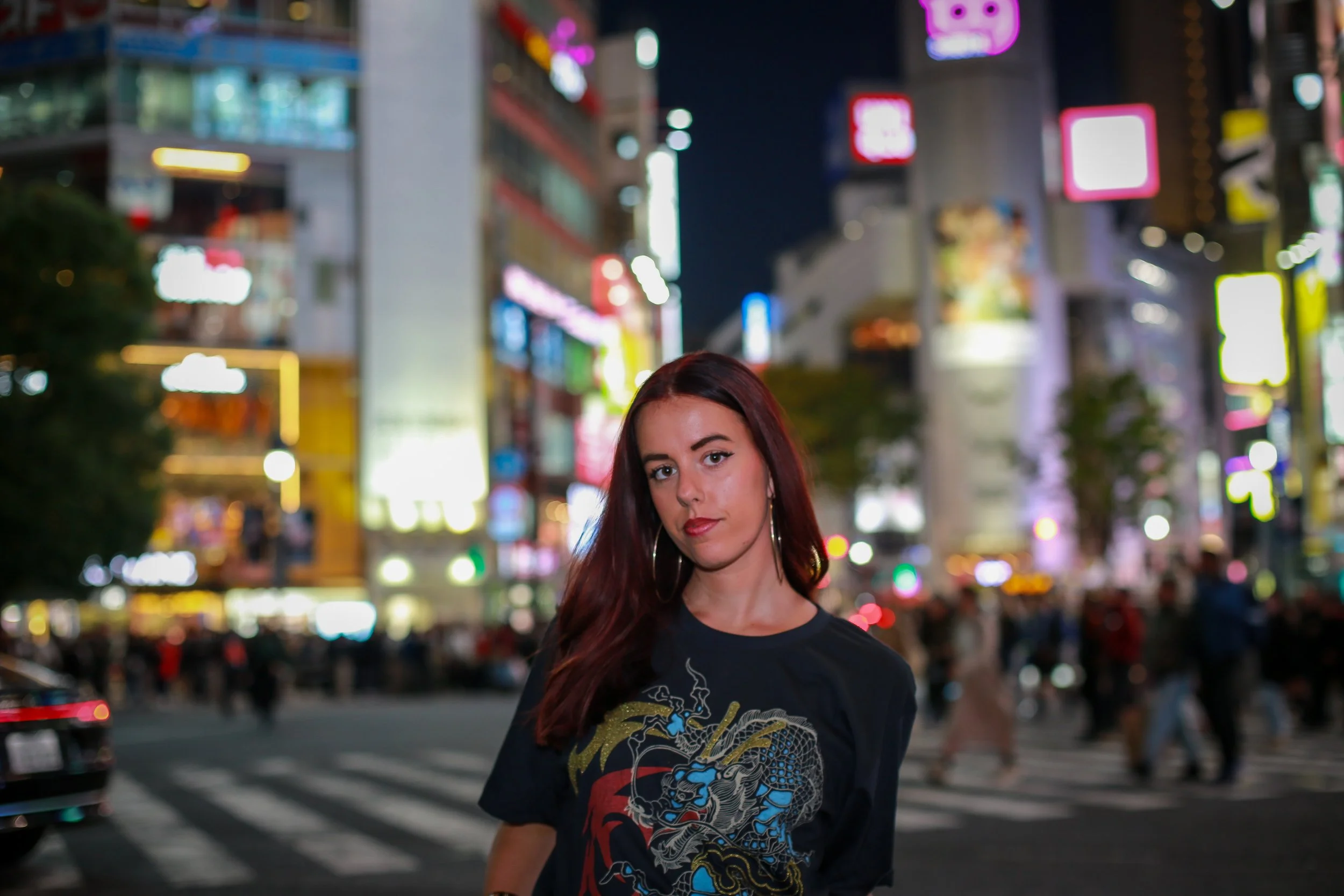A young woman with long red hair and hoop earrings standing on a city street at night, illuminated by colorful neon signs and busy traffic.