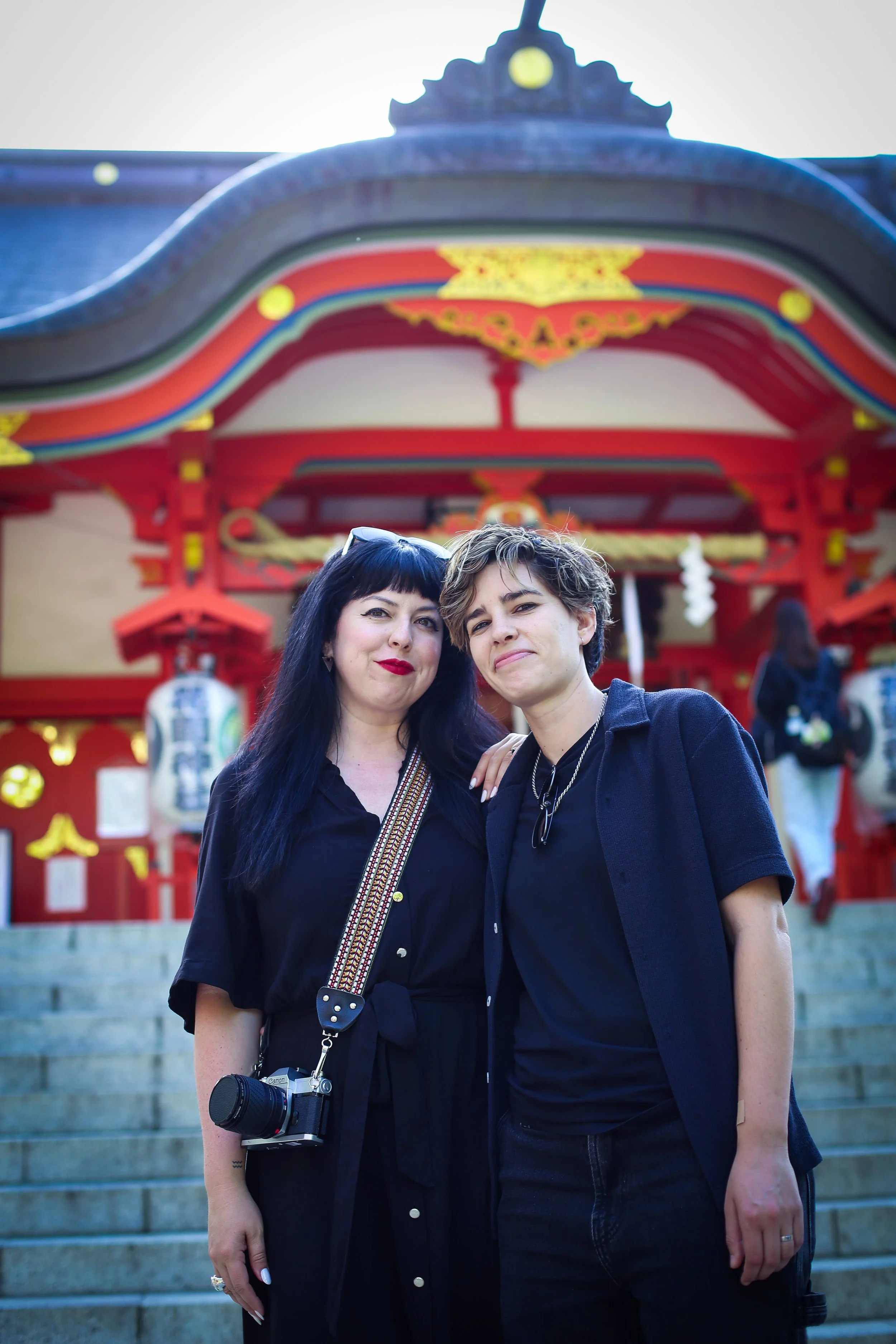 Two women standing on steps in front of a red and gold torii gate at a Japanese shrine, smiling at the camera.