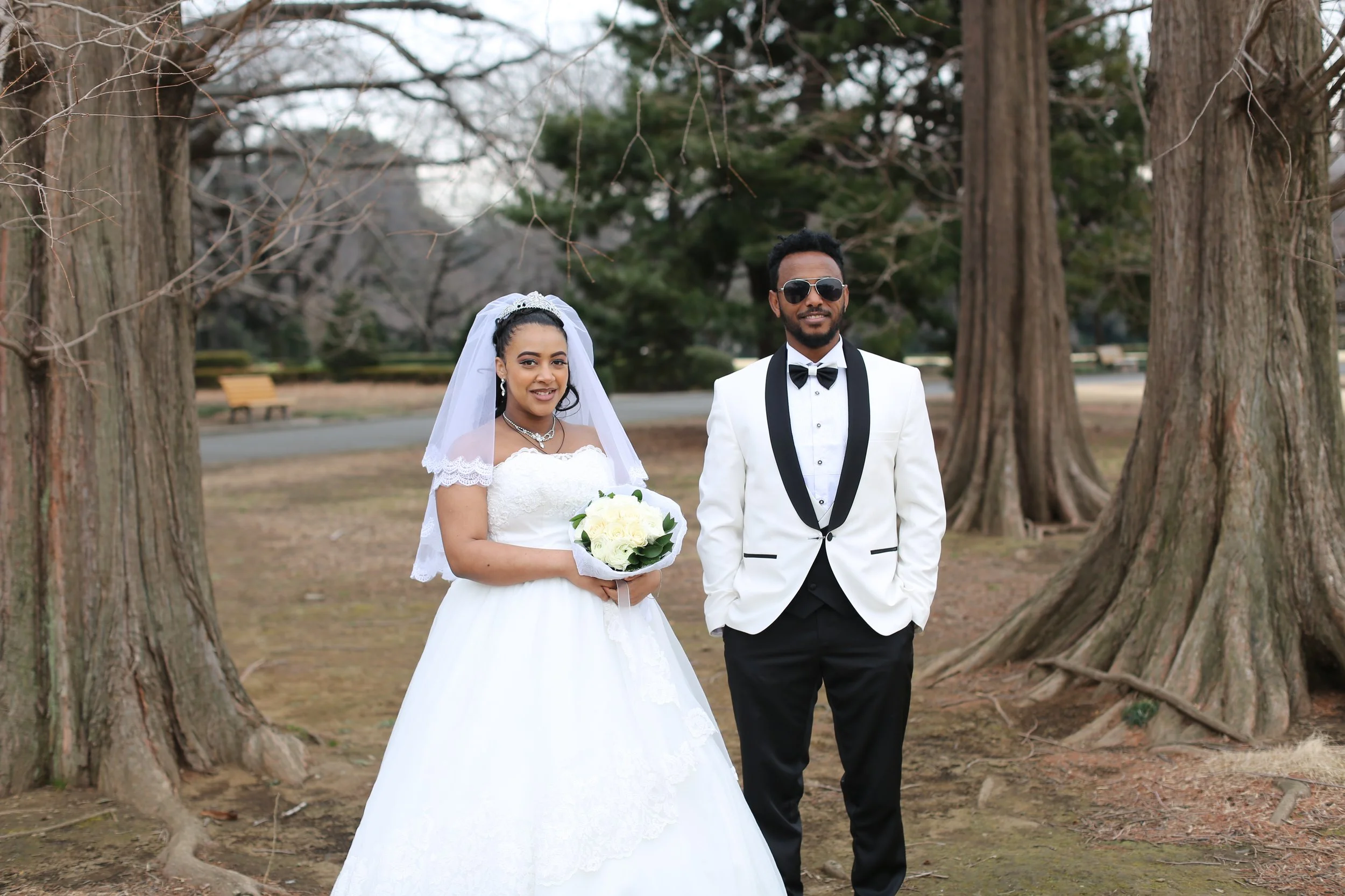A bride and groom standing outdoors between large trees in a park setting, smiling at the camera. The bride is holding a bouquet of white flowers and is dressed in a white wedding gown with a veil, while the groom is in a white tuxedo jacket with a b