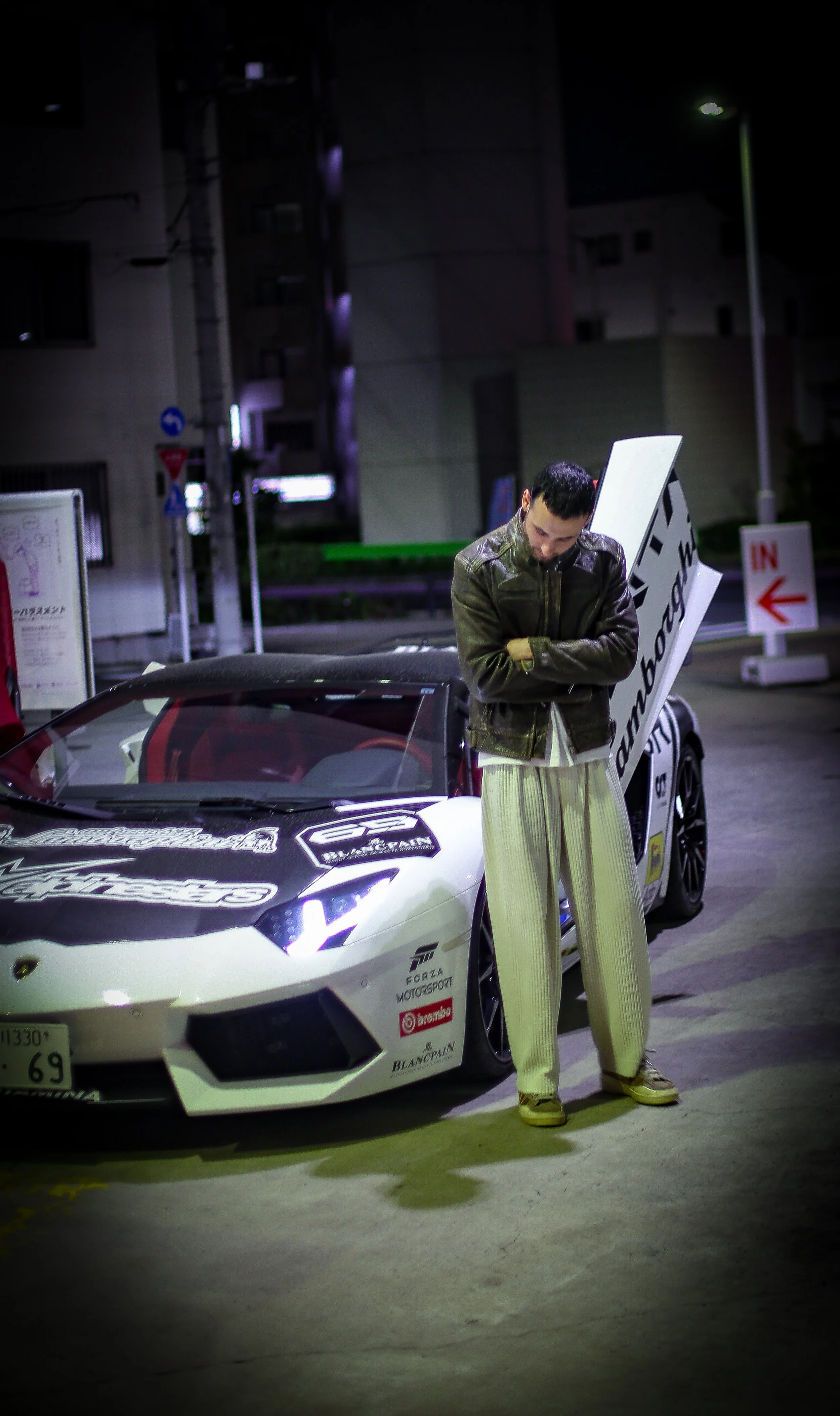A man wearing a leather jacket and cream-colored wide-legged pants is standing next to a white Lamborghini sports car at night. The car has various racing decals on it. The man is looking down with his arms crossed.