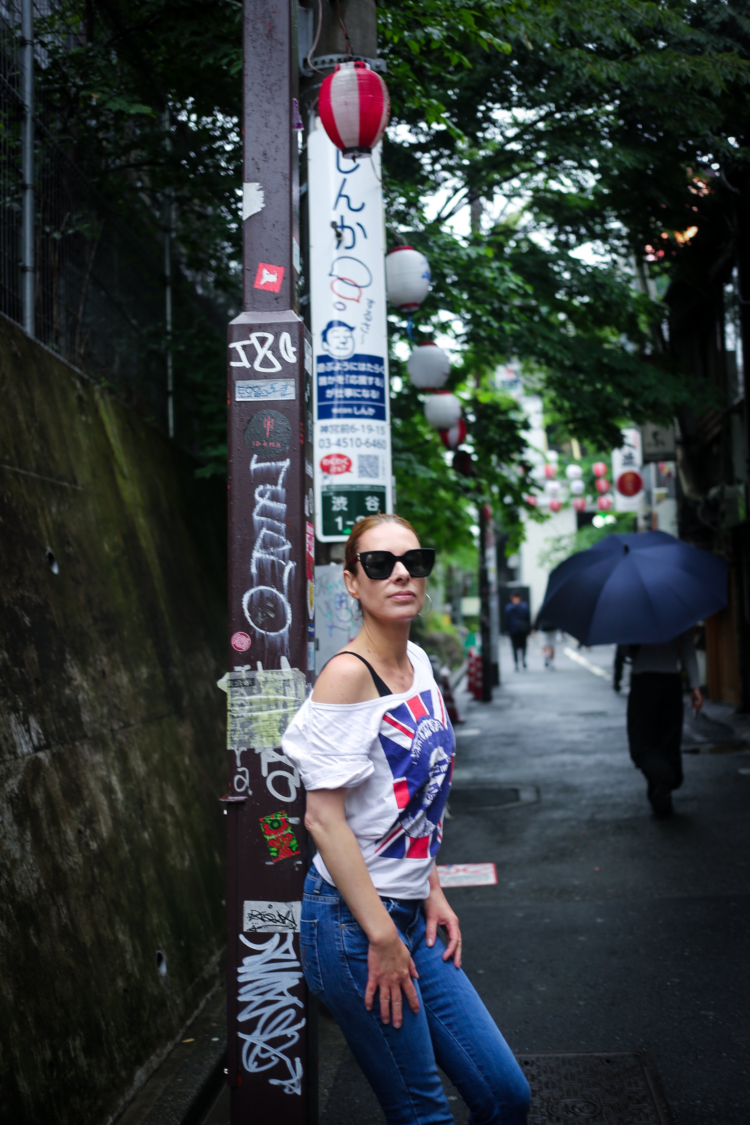 A woman wearing sunglasses and a T-shirt with a Union Jack design, standing on a city street beside a pole covered in graffiti and stickers, with trees and buildings in the background. She is holding a small bag and appears to be posing for the photo