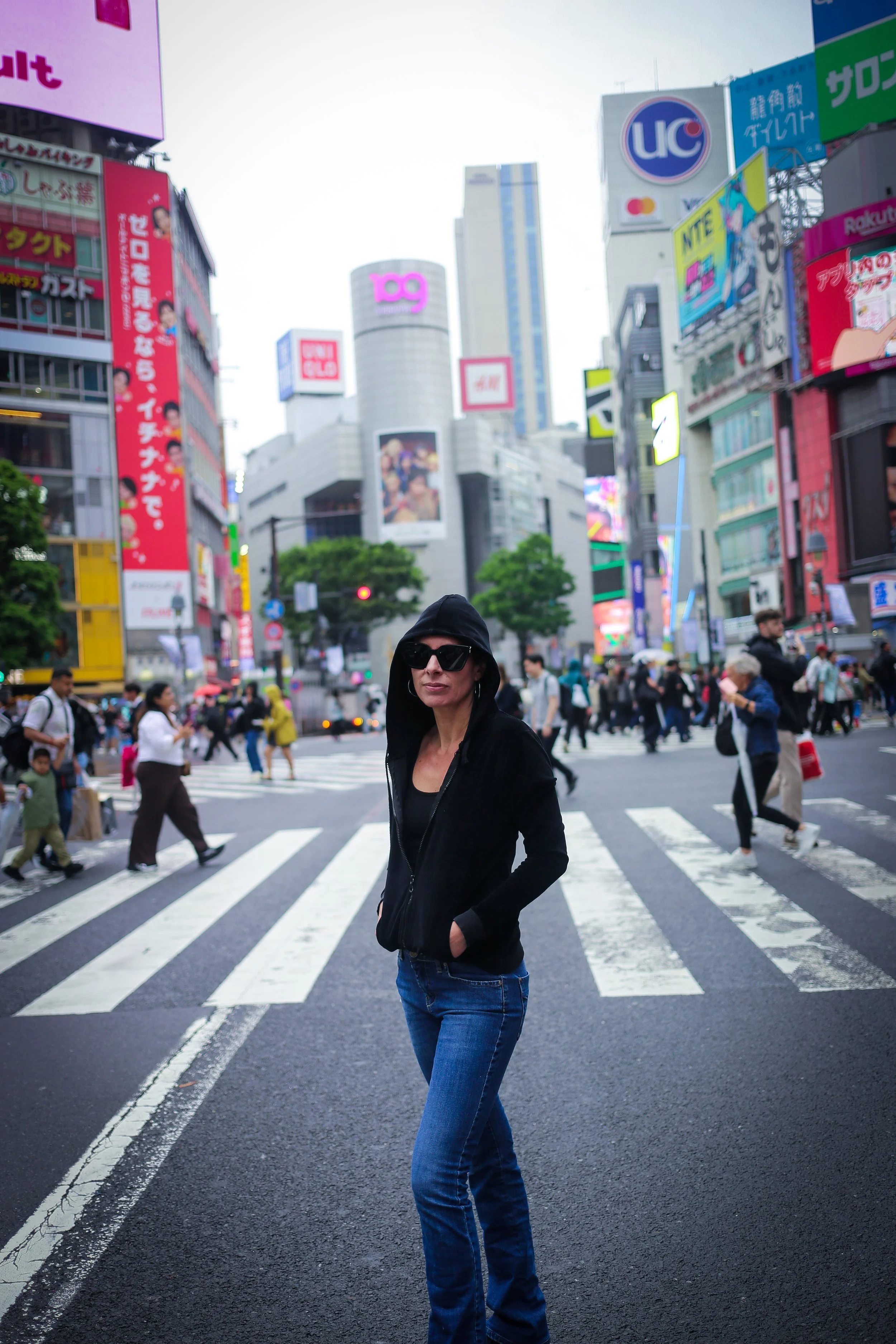 A woman in black hoodie and sunglasses standing on a crosswalk in a busy city with tall buildings and neon signs.