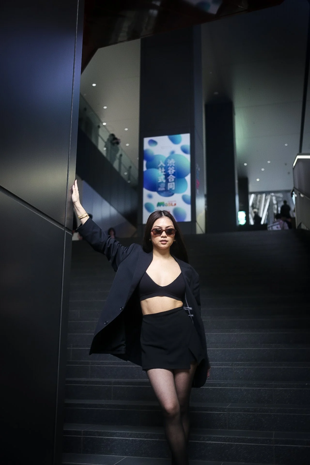 A woman wearing sunglasses, a black blazer, a black crop top, a black skirt, and black fishnet stockings, standing on dark stairs in a modern building with a large digital advertisement in the background