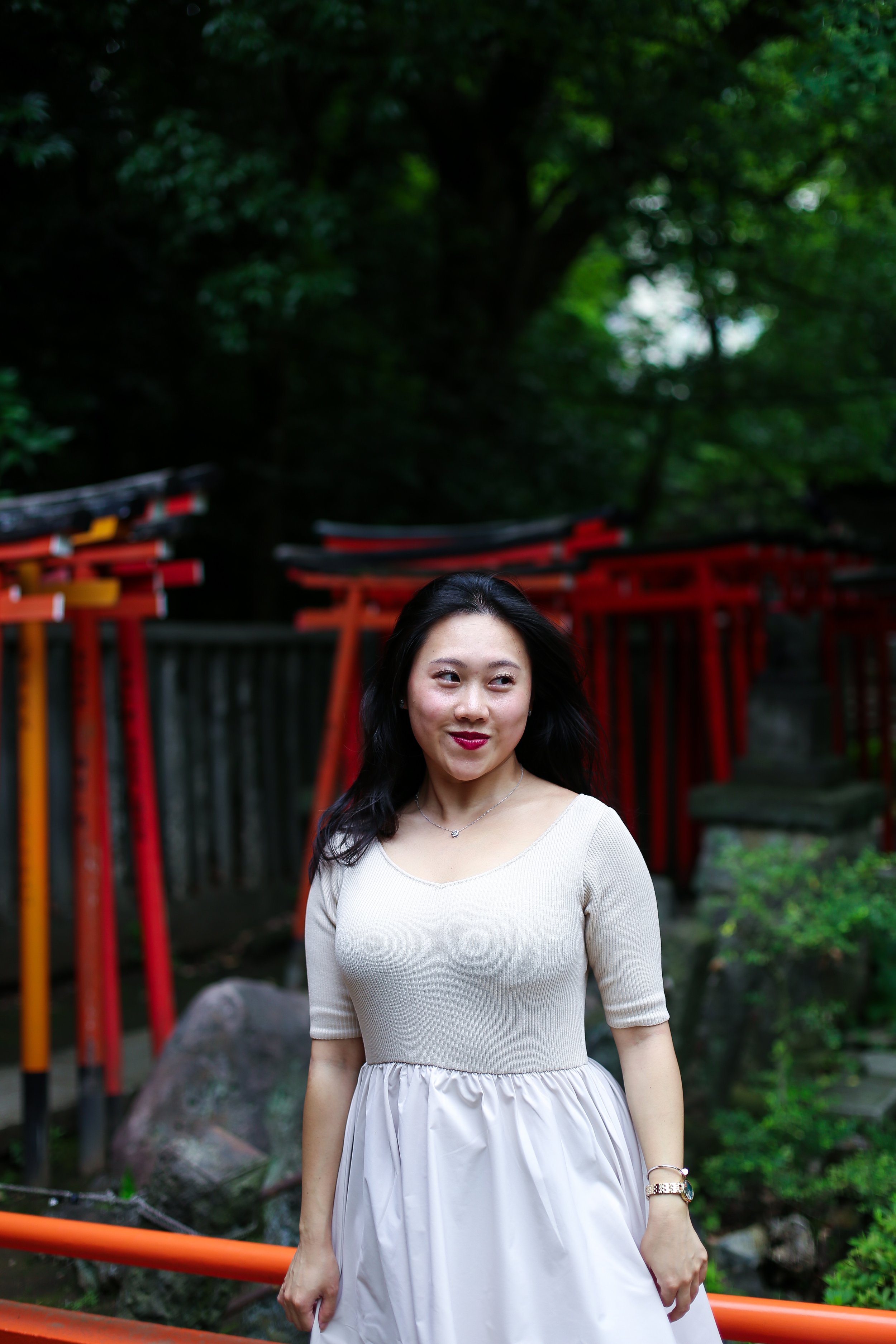 A woman with long black hair stands outdoors near a rock and orange railing, with red bridge structures and green trees in the background.