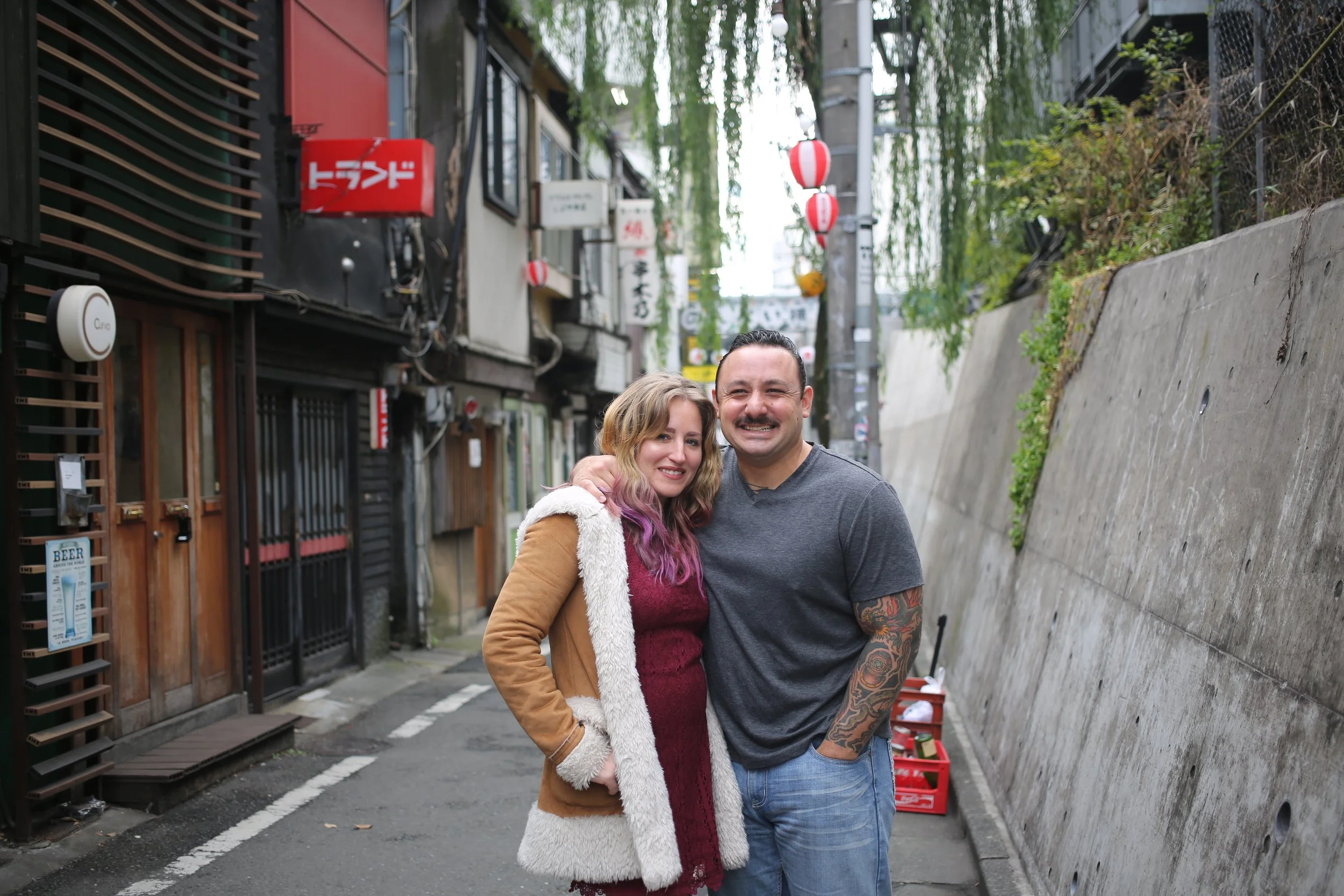 A smiling man and woman standing together outdoors on a narrow street with traditional Japanese lanterns hanging above. The woman has purple-tinted hair and is wearing a brown jacket with white fur trim, while the man has tattoos on his arms and is w