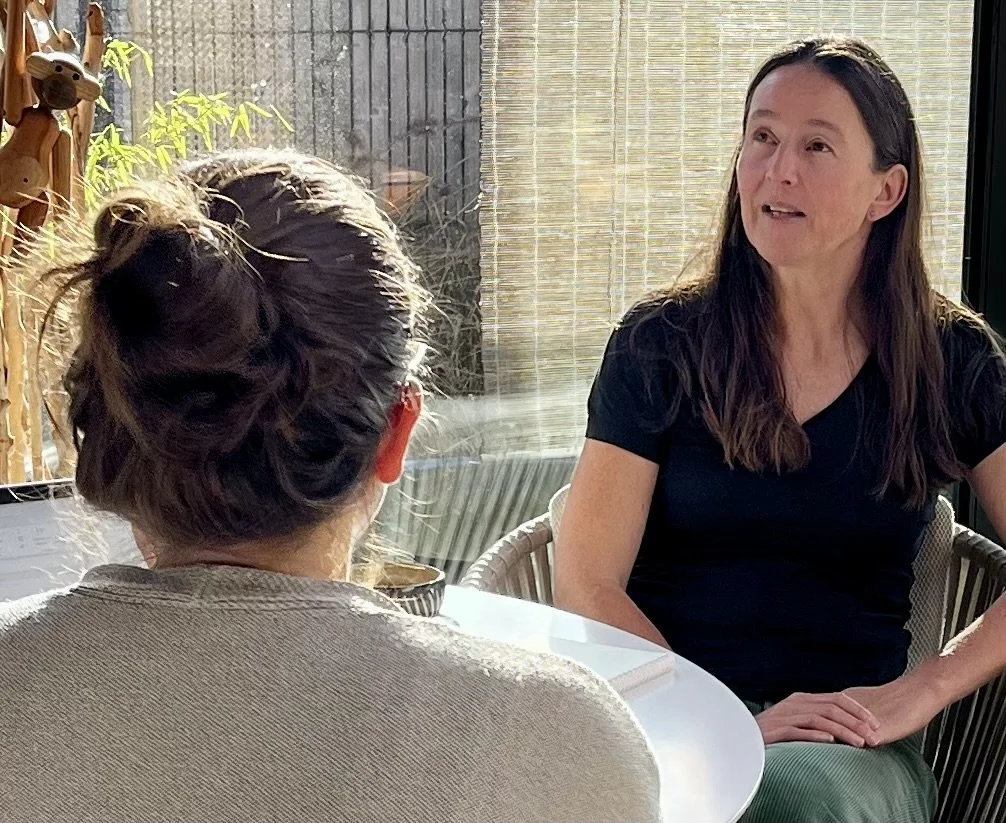 Twee vrouwen die in een gesprek zitten, één van hen heeft haar rug naar de camera en de ander kijkt naar haar, binnen in een kamer met veel natuurlijk licht.