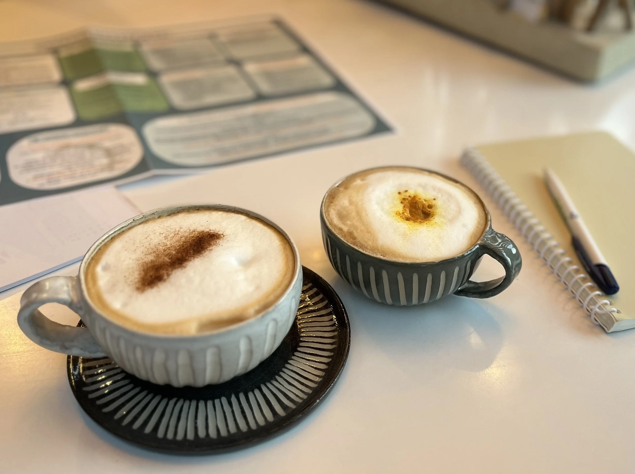 Twee koppen met cappuccino coffee op een witte tafel, daarnaast notitieboekje en pen, de ondernemersscan op de achtergrond.