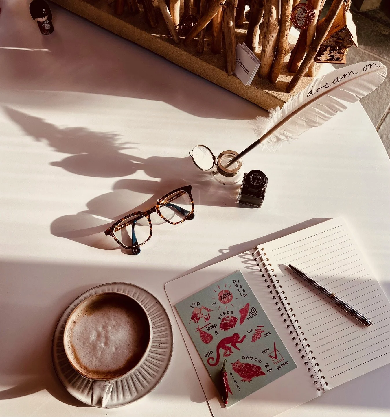Een tafel met een kop koffie, een paar leesbrillen, een open notitieboek en een pen, met daarnaast een glazen inktpot en een veer met 'dream on' opgeschreven. Op de achtergrond een rij takken en kleine decoraties.