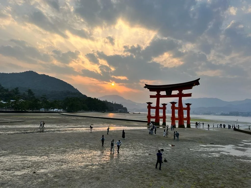 Een grote rode torii-poort op het strand met mensen die eromheen lopen, bij zonsondergang met bergen op de achtergrond en gedeeltelijk bewolkte hemel.
