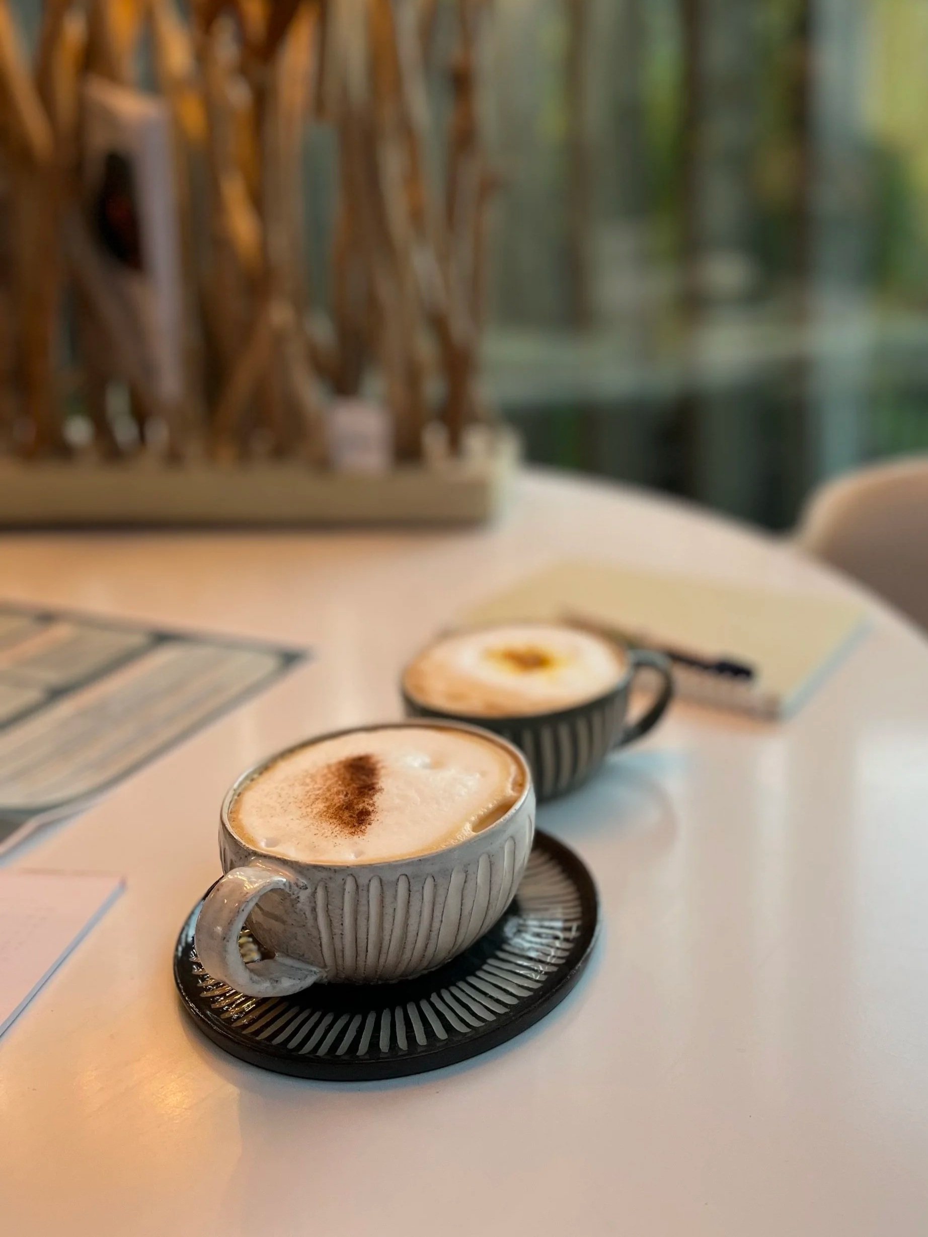 Twee kopjes cappuccino op een witte tafel in een café, met een boek en decoratief huisje op de achtergrond.